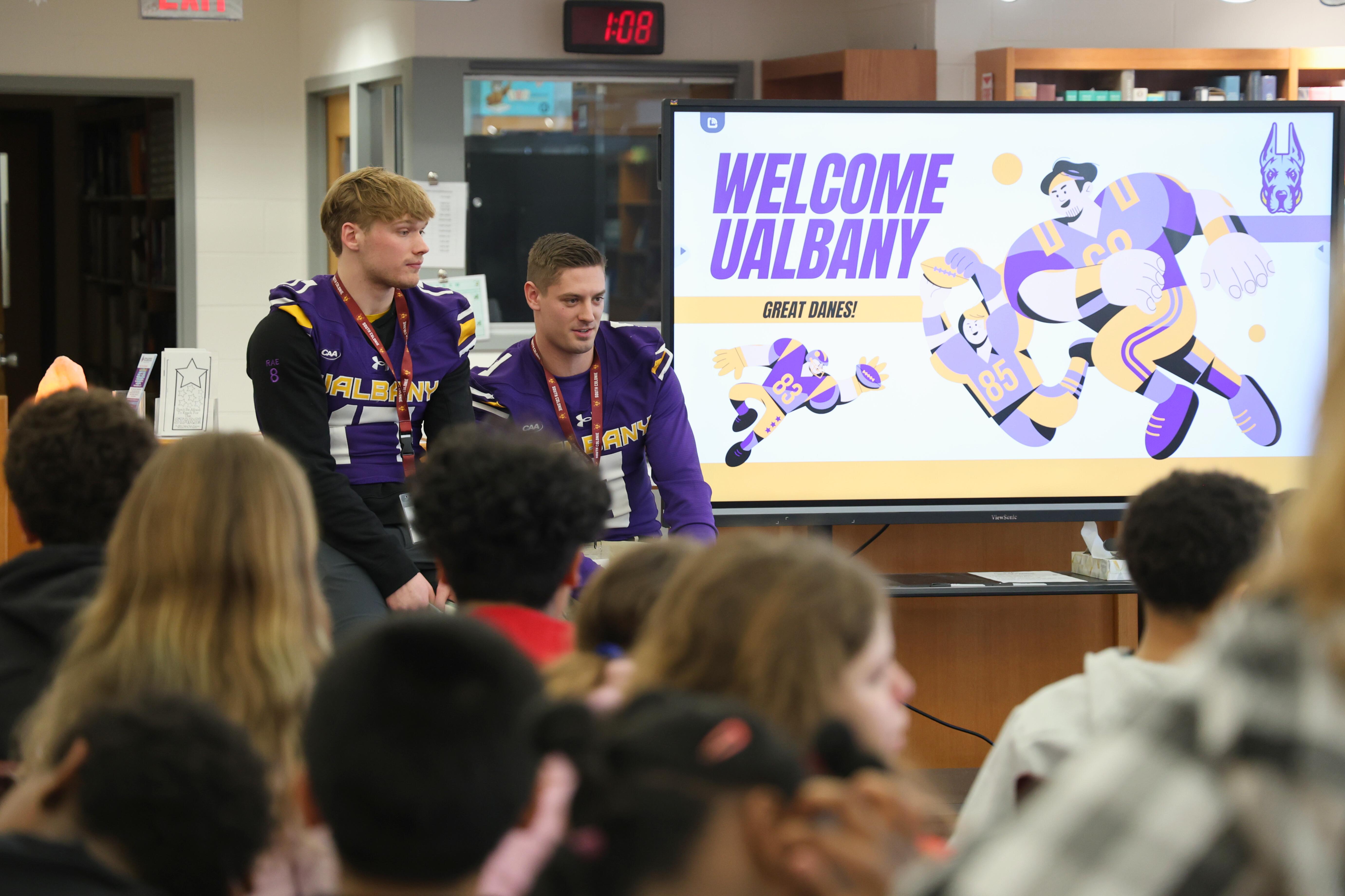 Two people in purple and gold jerseys stand in front of a screen that says 'Welcome UAlbany'.