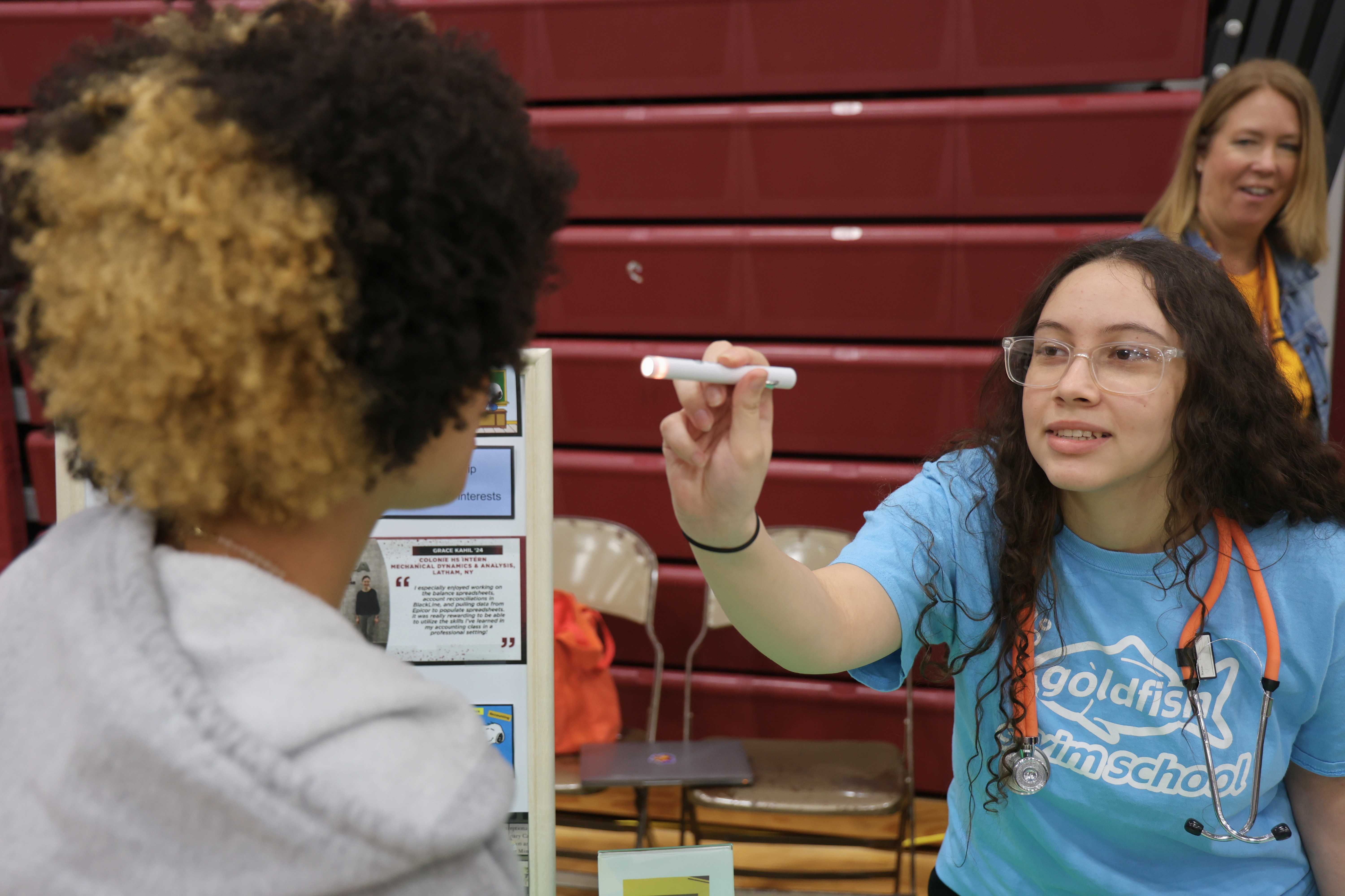 A student wearing a stethoscope shines a small flashlight into another student's eye