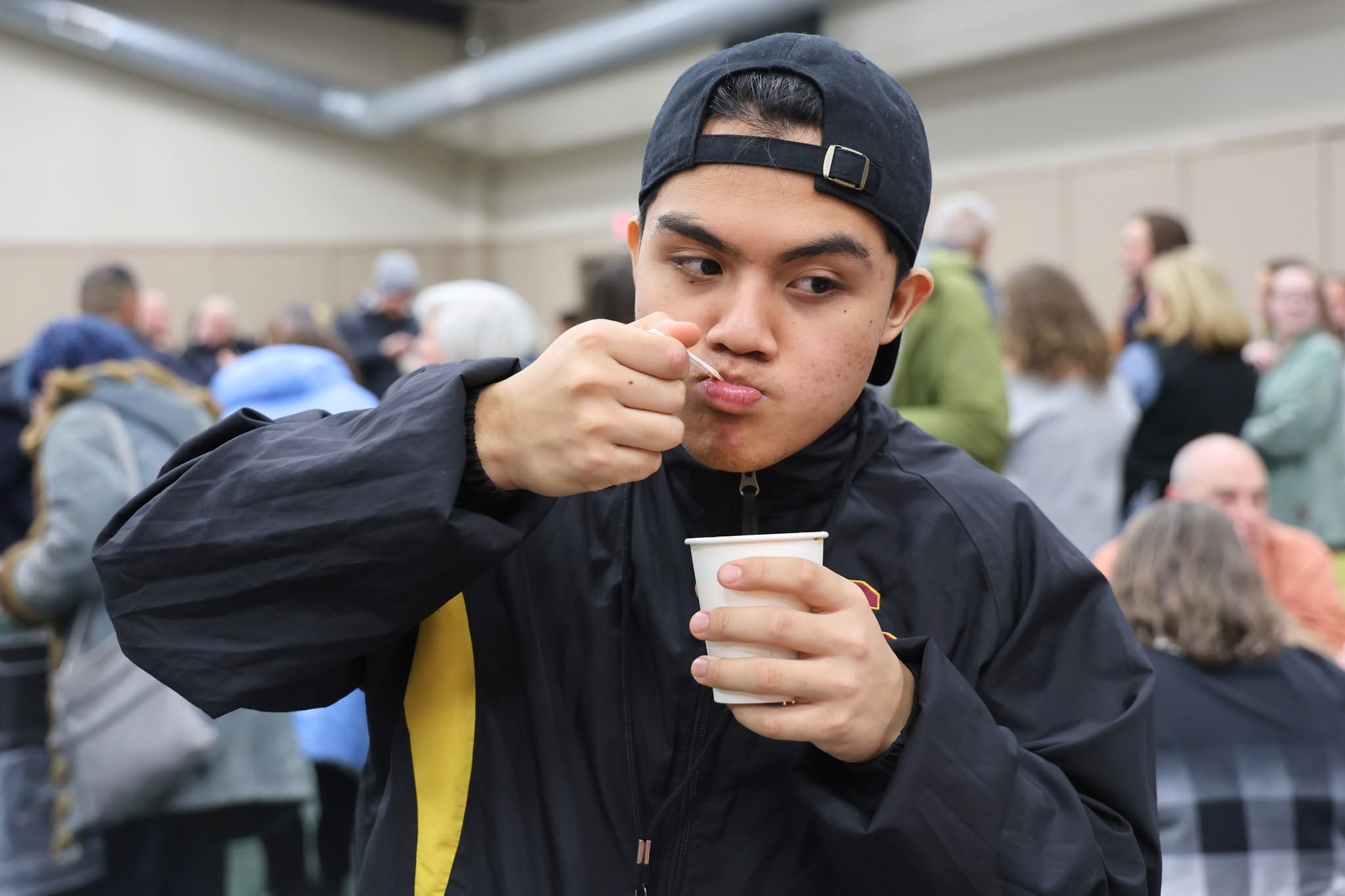 A young person in a black baseball cap and jacket eats from a small white cup.