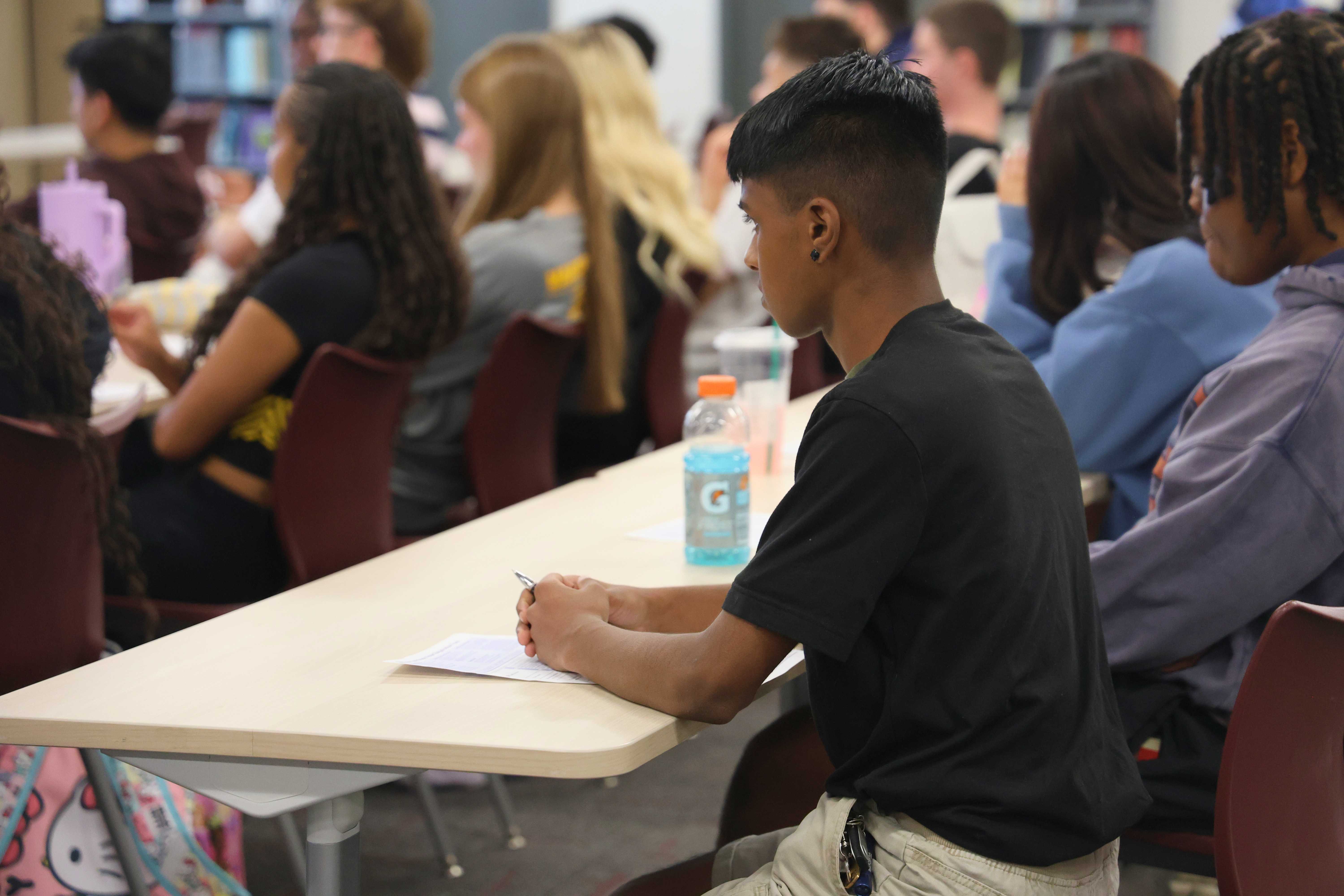 A group of students sits attentively at desks in a well-lit classroom.