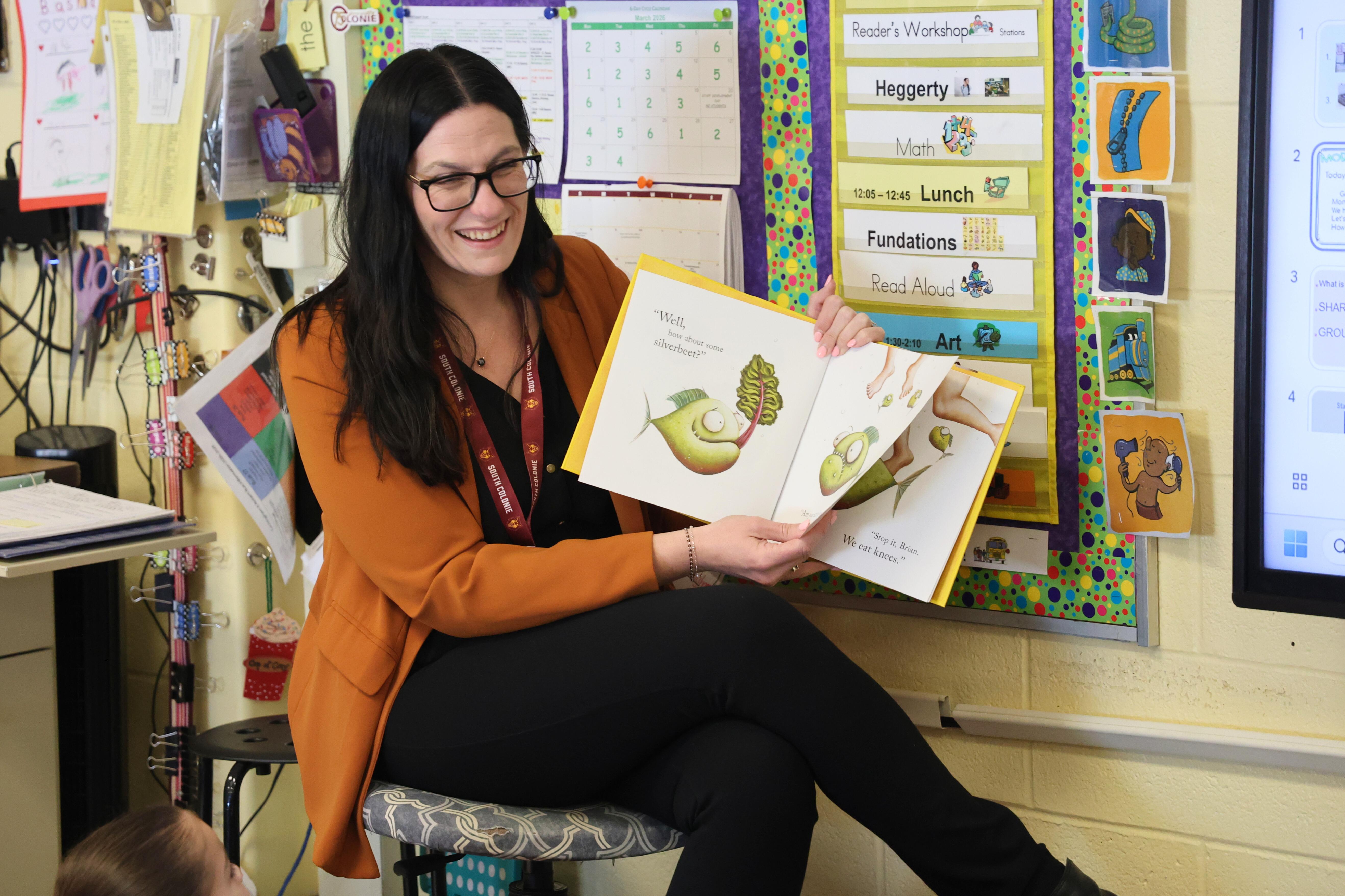 A teacher smiles while reading a picture book to a class.