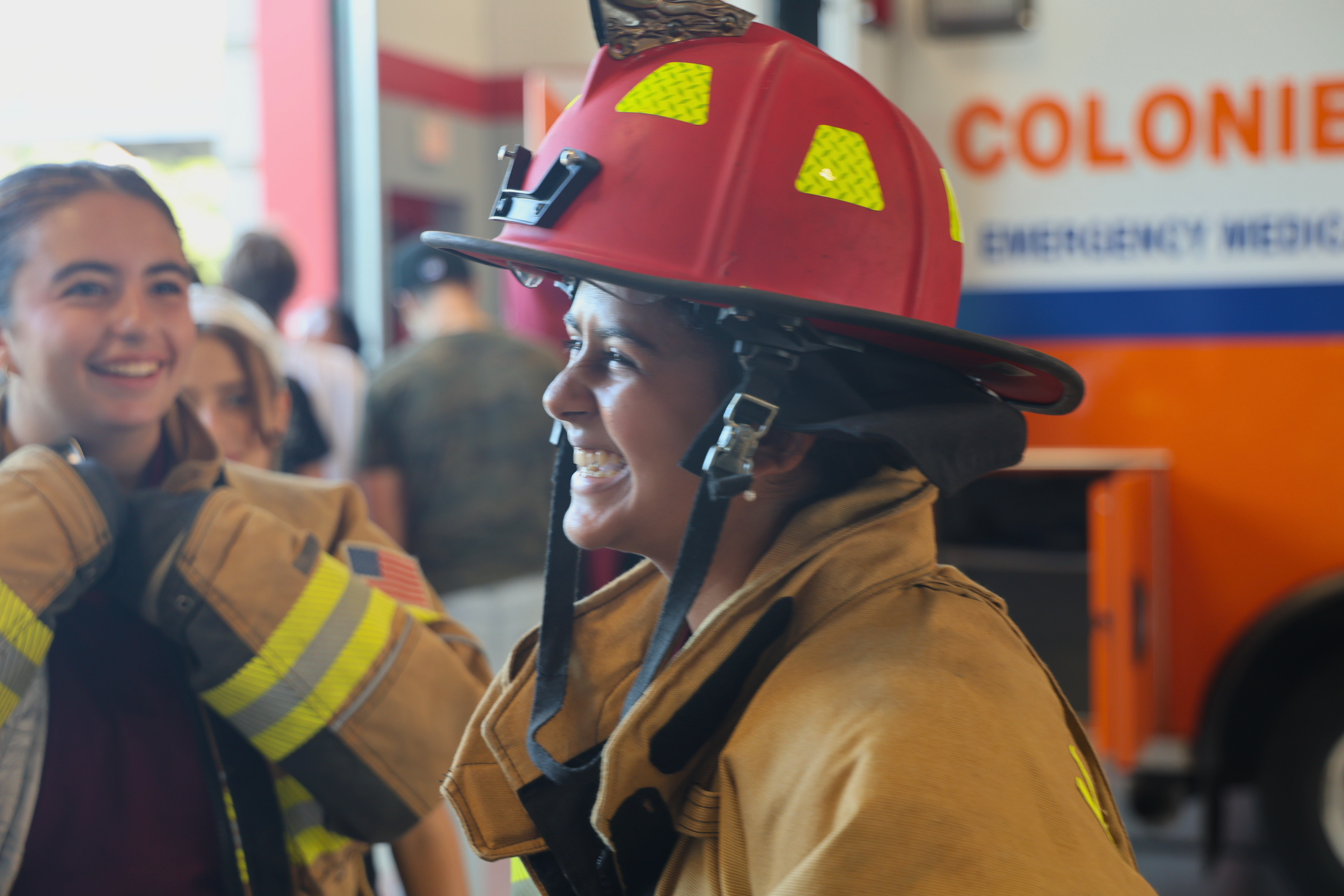 A person wearing a large red firefighter helmet and tan turnout gear smiles inside a fire station.