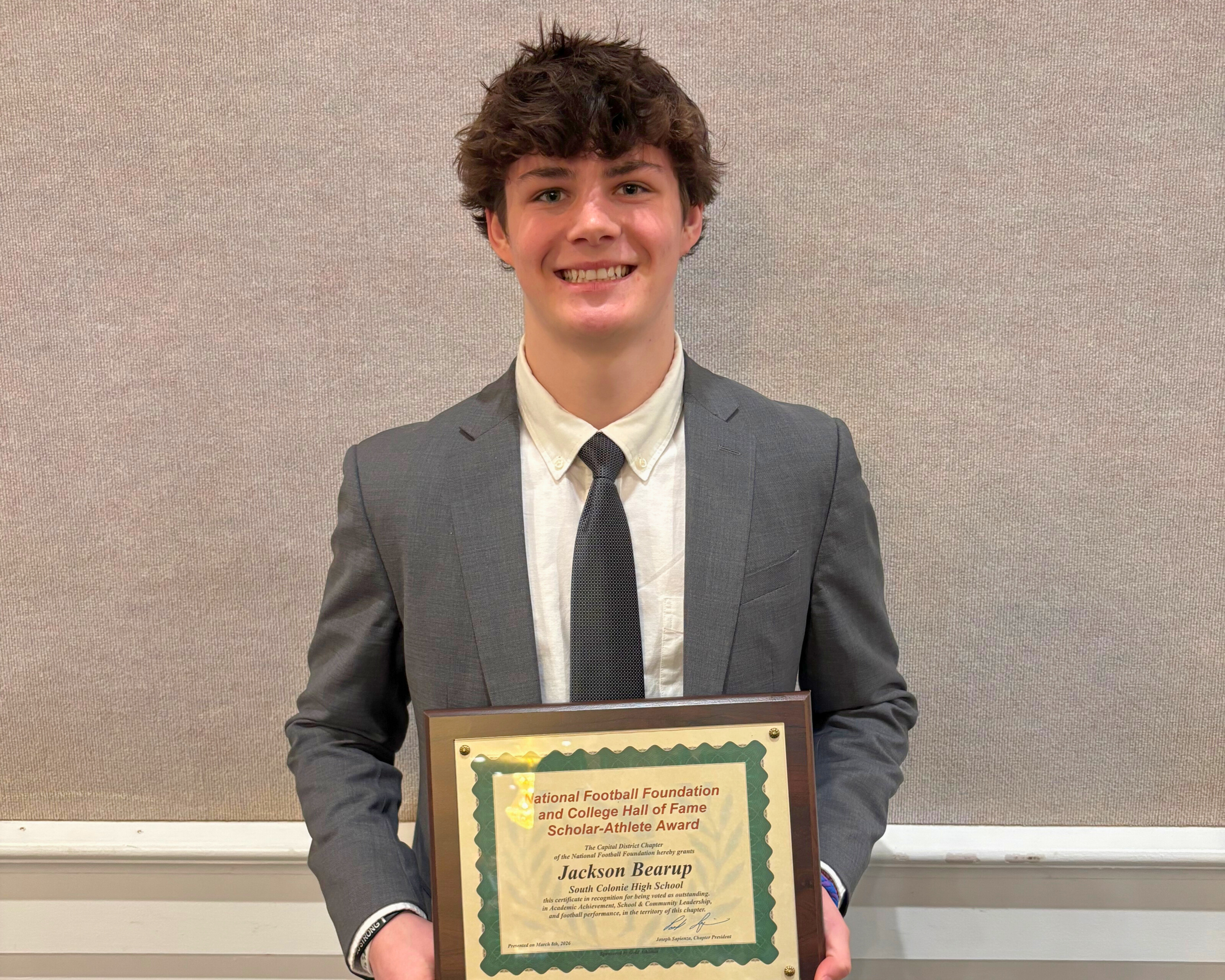 A young man in a suit smiles, holding an award plaque.
