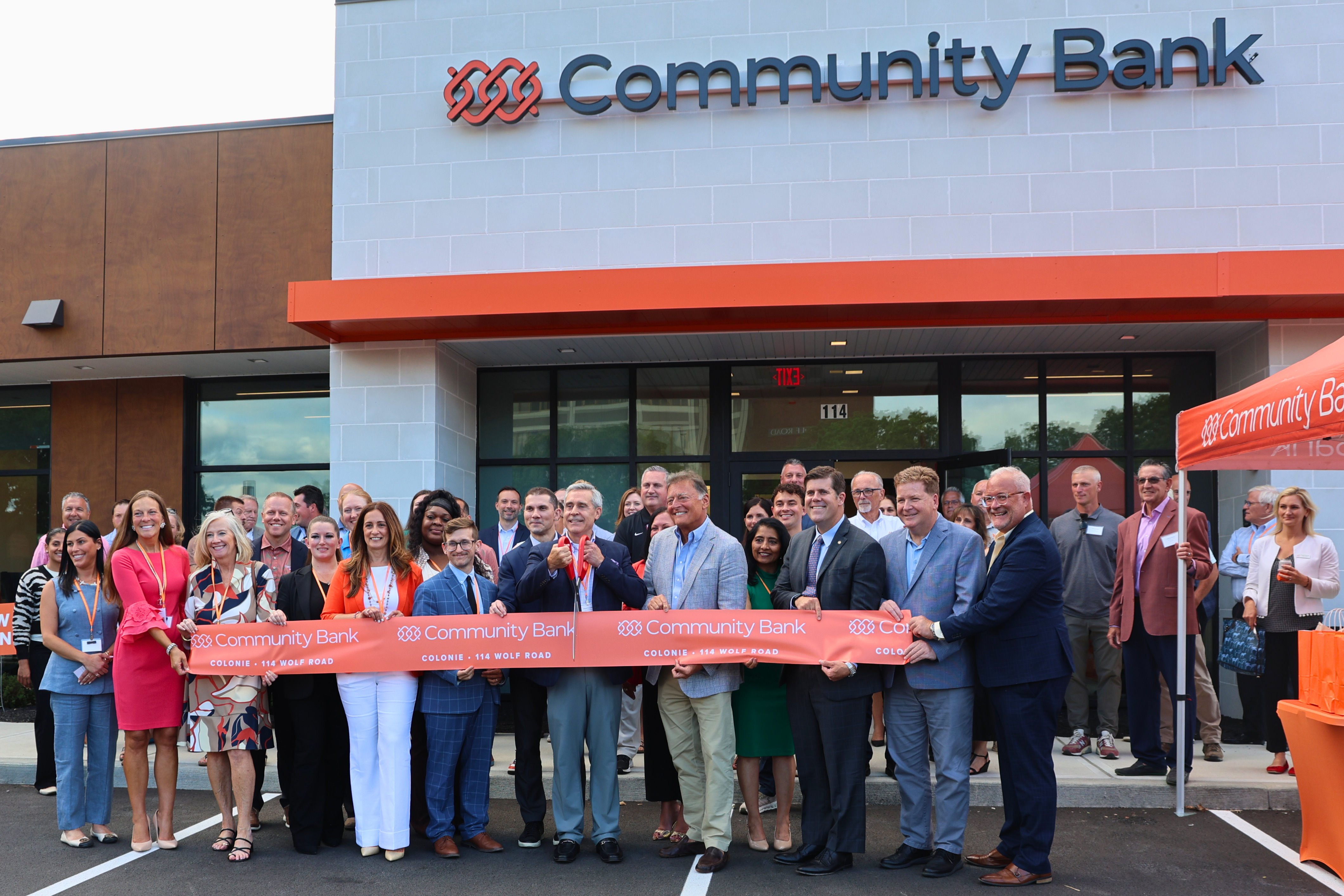 A group of people in business attire hold a large orange ribbon for a ribbon-cutting ceremony in front of a Community Bank building.