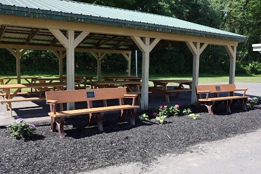 An open-air pavilion with wooden picnic tables and benches, set against a backdrop of trees.