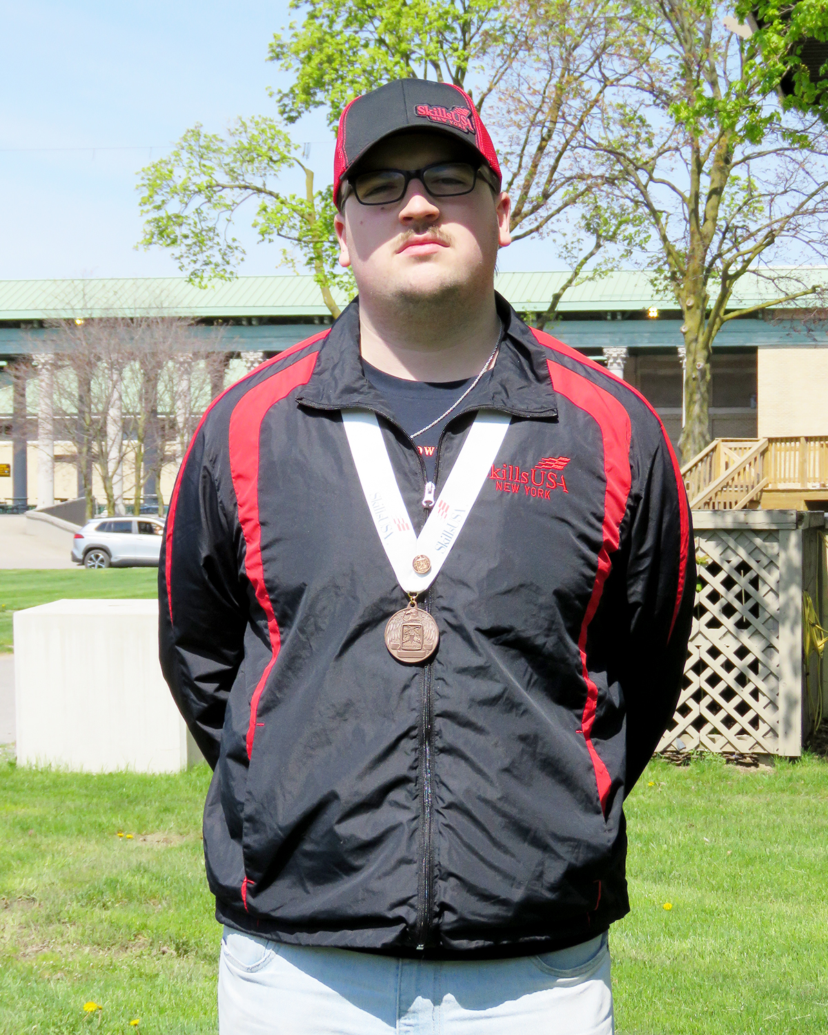 A student wearing a black and red jacket and a bronze medal stands outdoors.