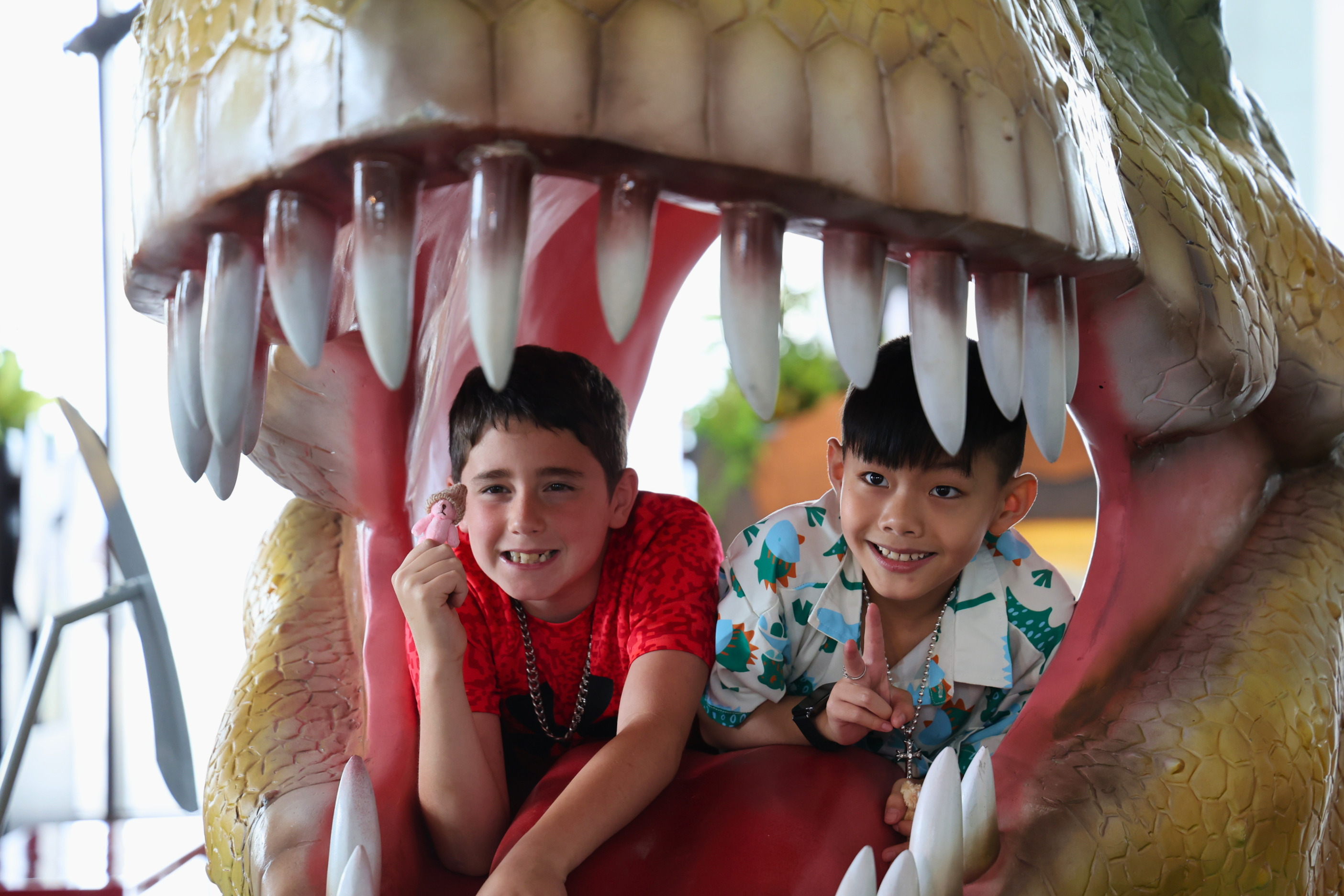 Two smiling boys peek out from the mouth of a large, colorful dinosaur prop.