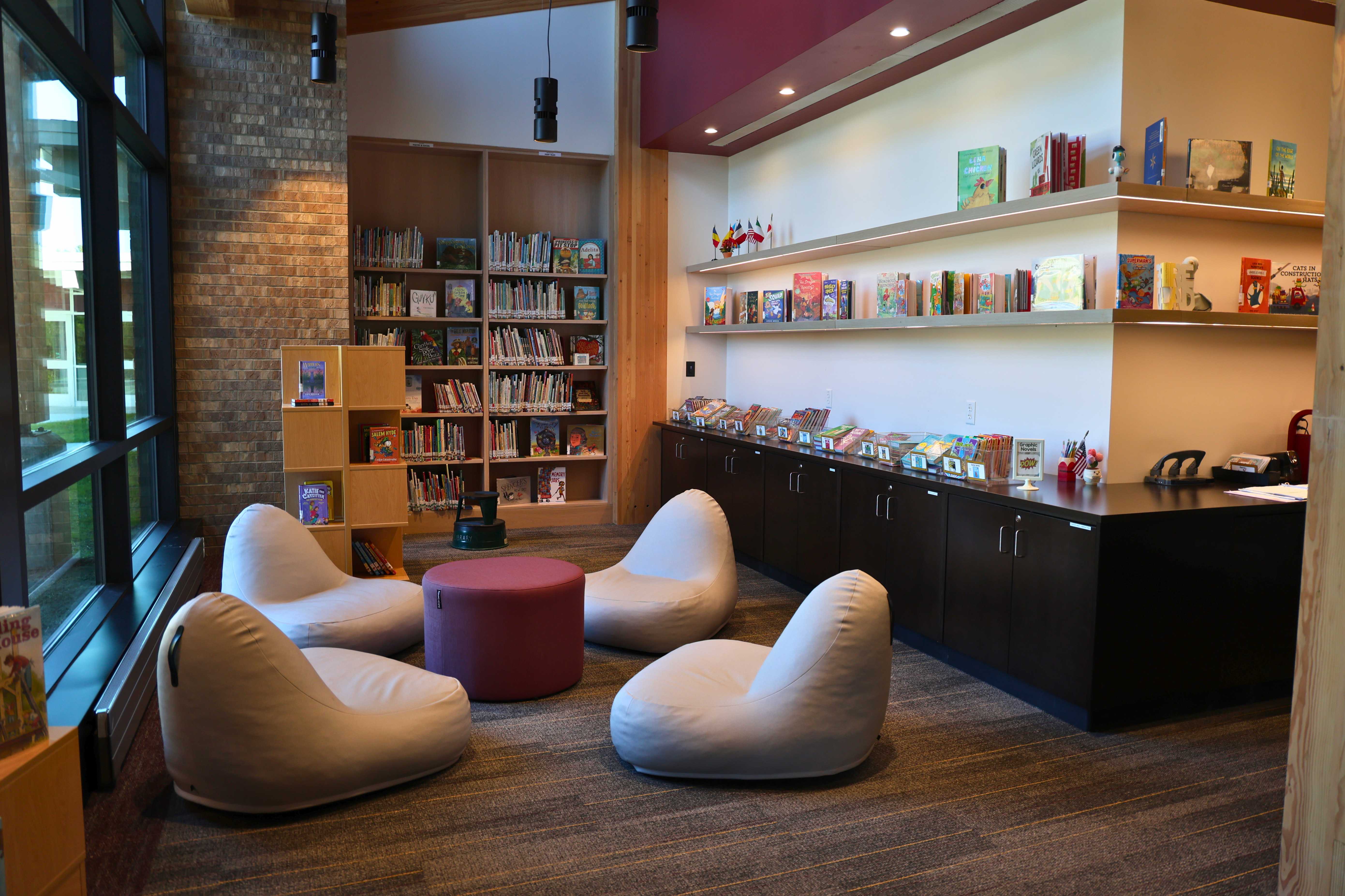 A cozy seating area with four white beanbag chairs and a red round ottoman inside a well-lit library.