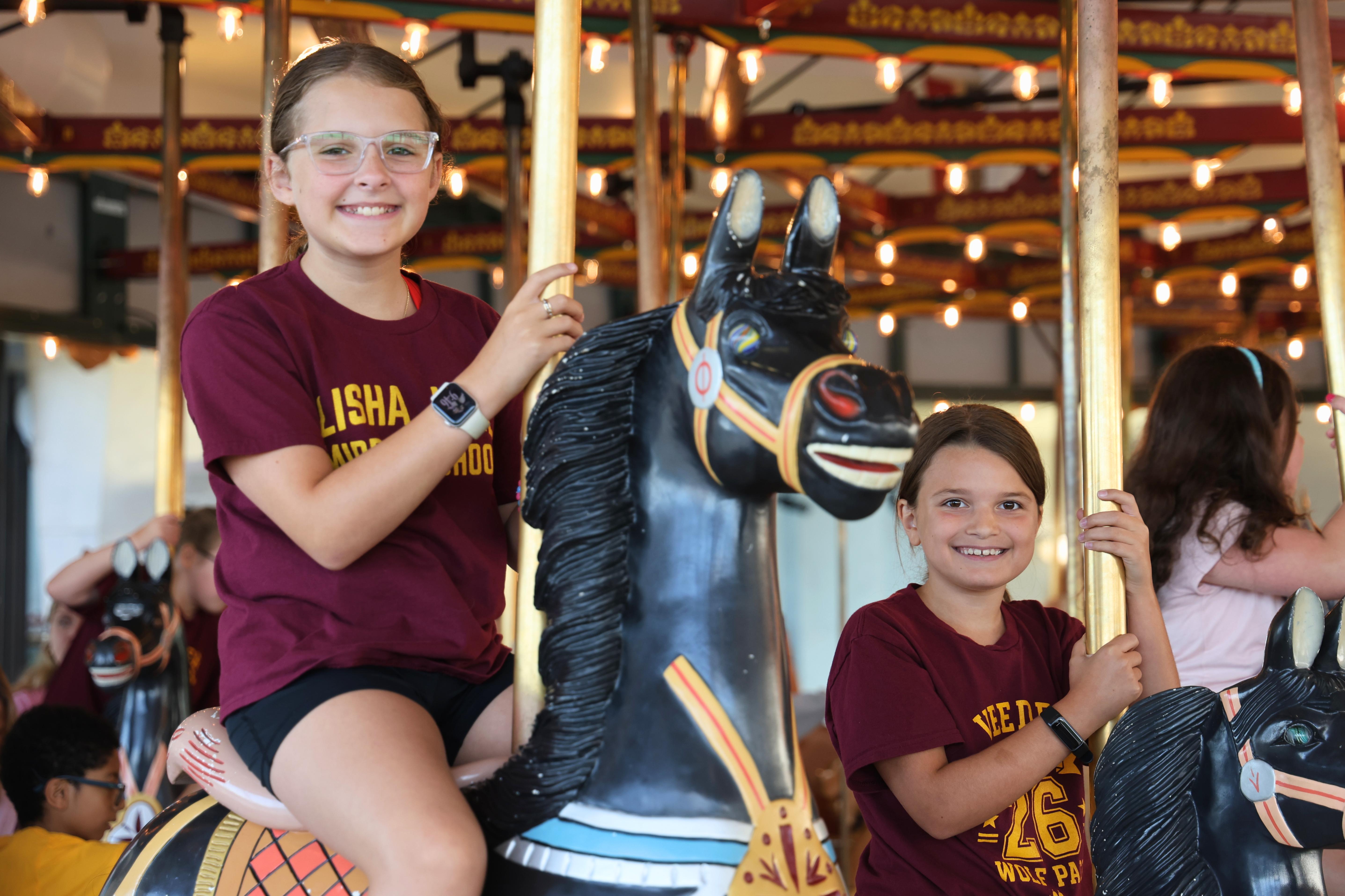 Two smiling girls in maroon shirts ride carousel horses, holding onto the poles.