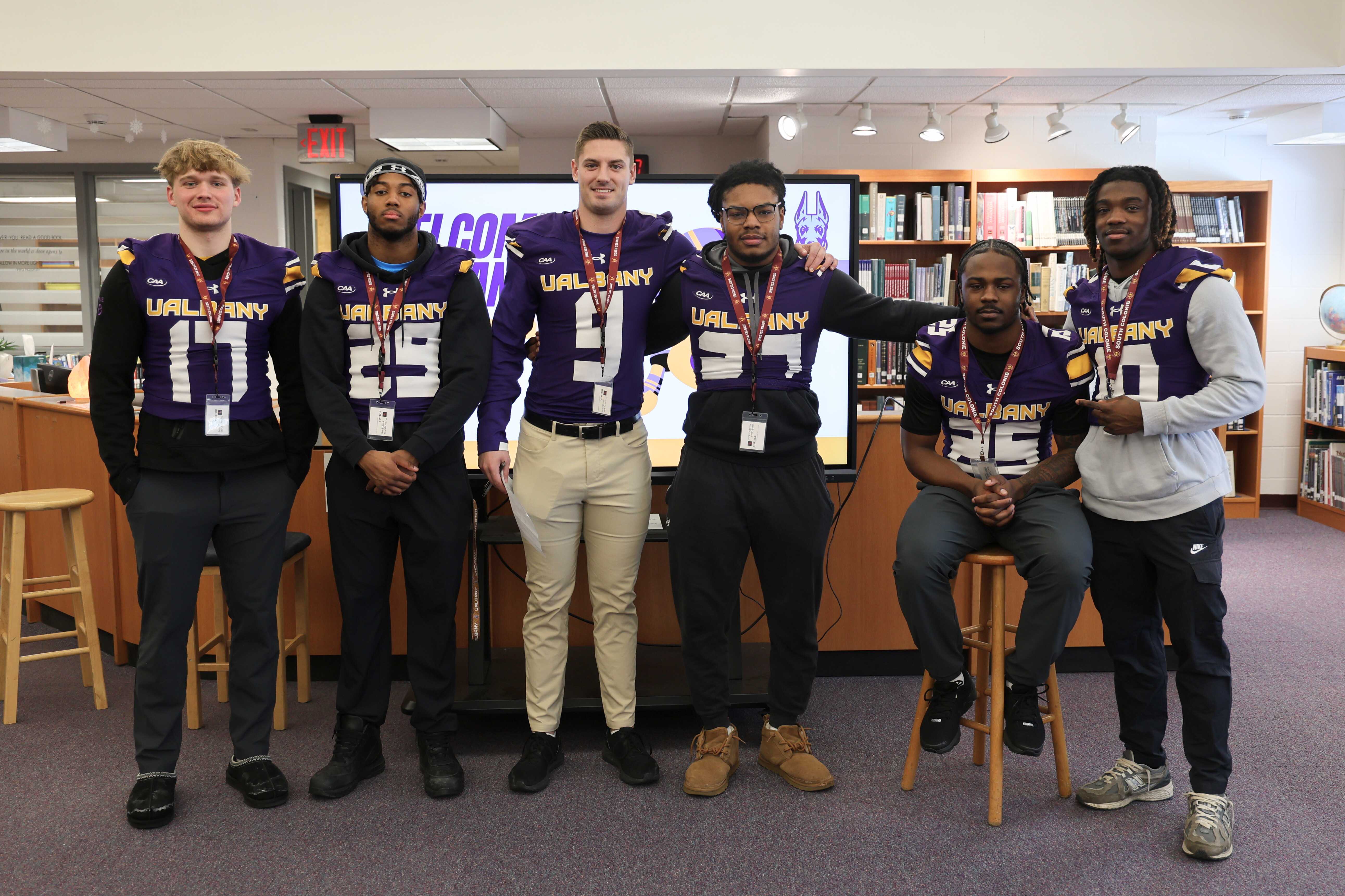 A group of six UAlbany football players pose for a photo in a library setting.