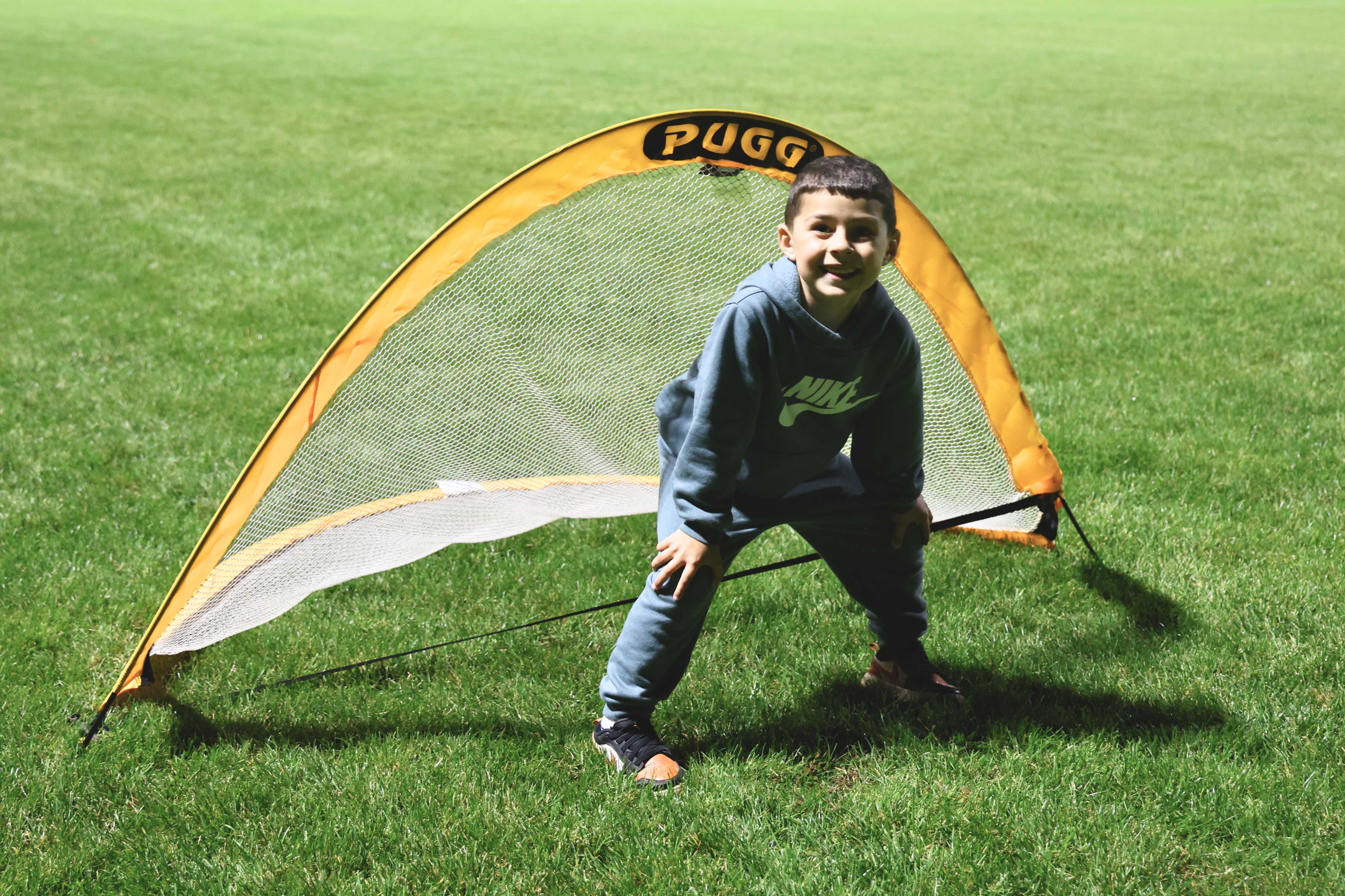 A child plays as a goalkeeper in front of a small orange and white pop-up soccer net on a grassy field.