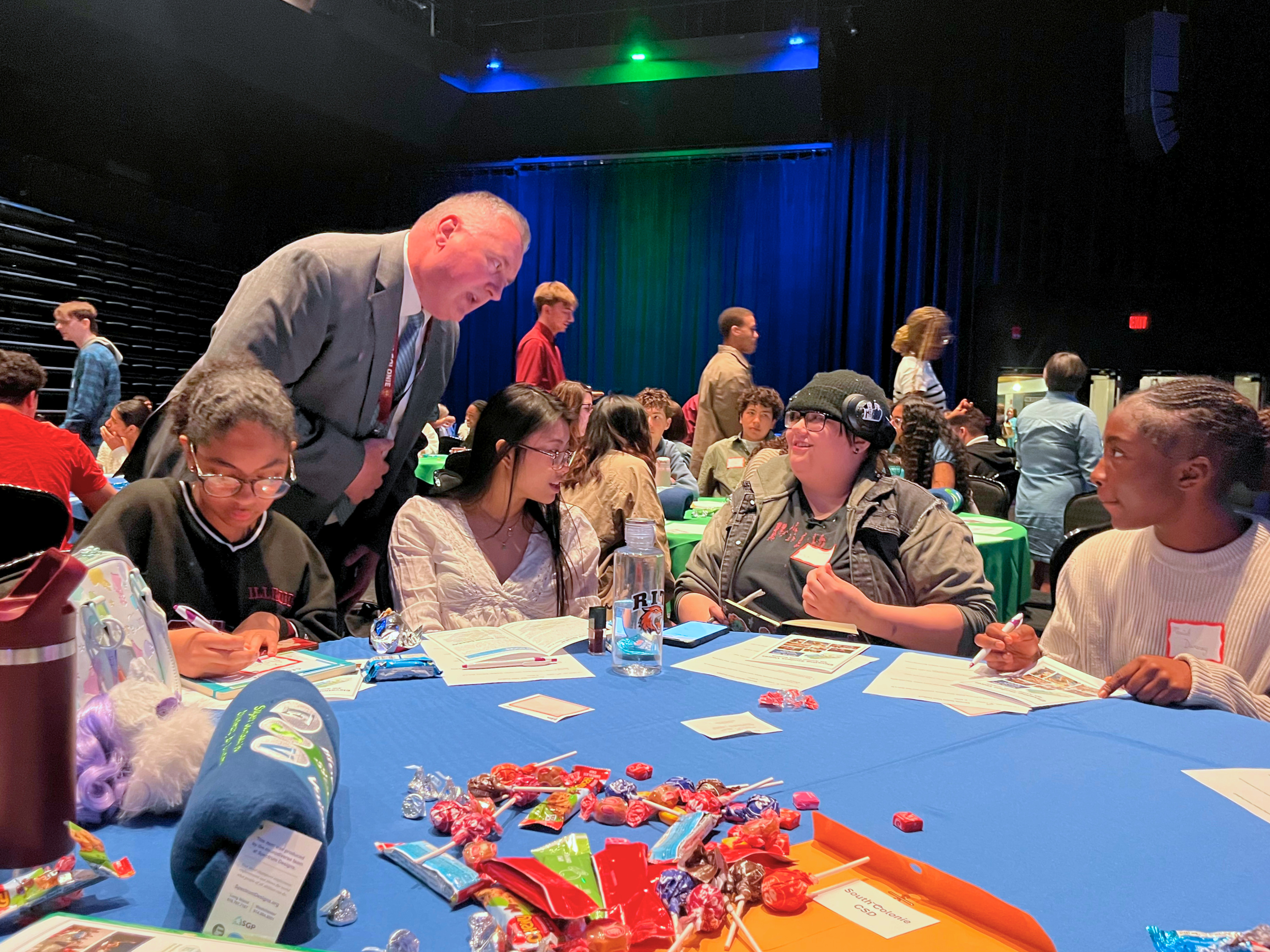 People sit at tables, engaged in an activity, with a man in a suit speaking to students.