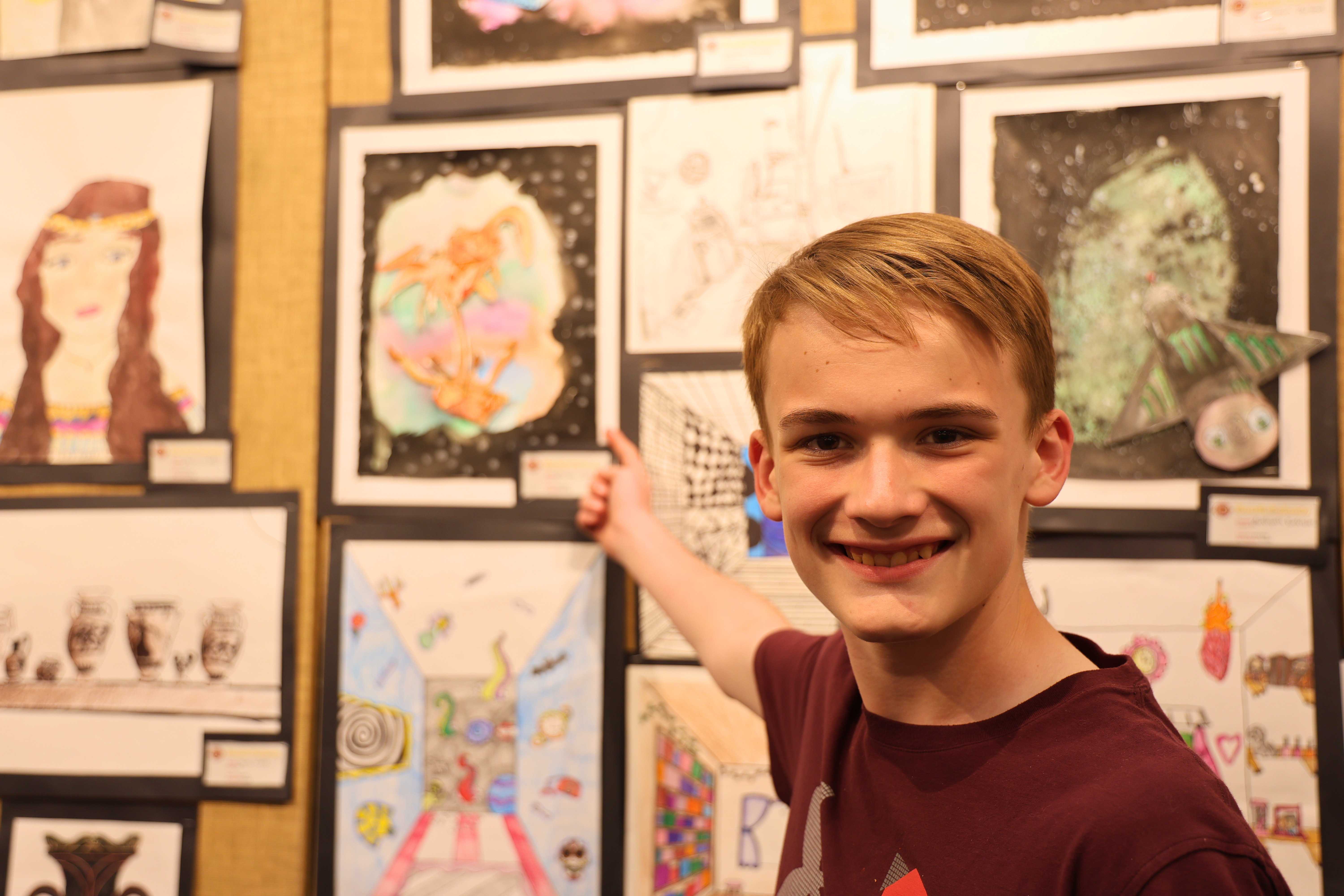 A young person smiles while pointing at artwork displayed on a wall.