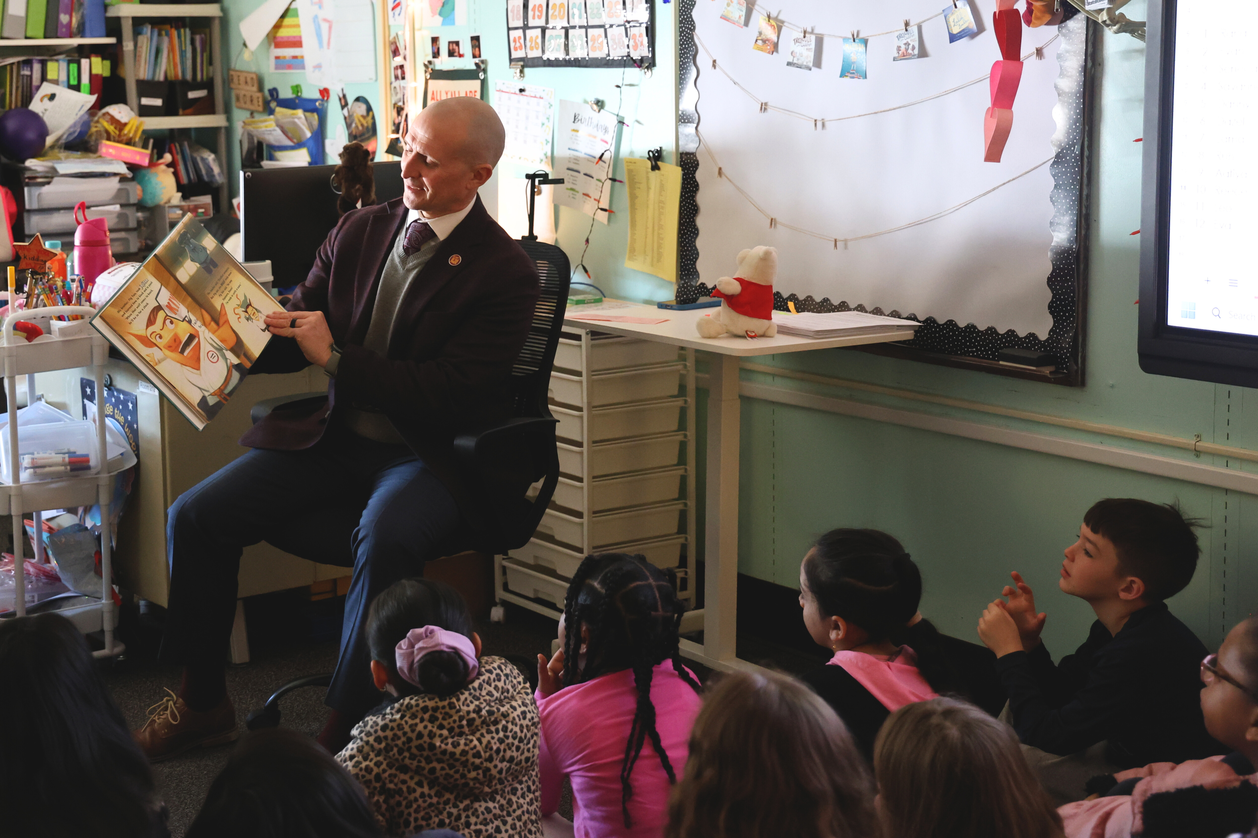 A man reads a book to a group of children in a classroom setting.