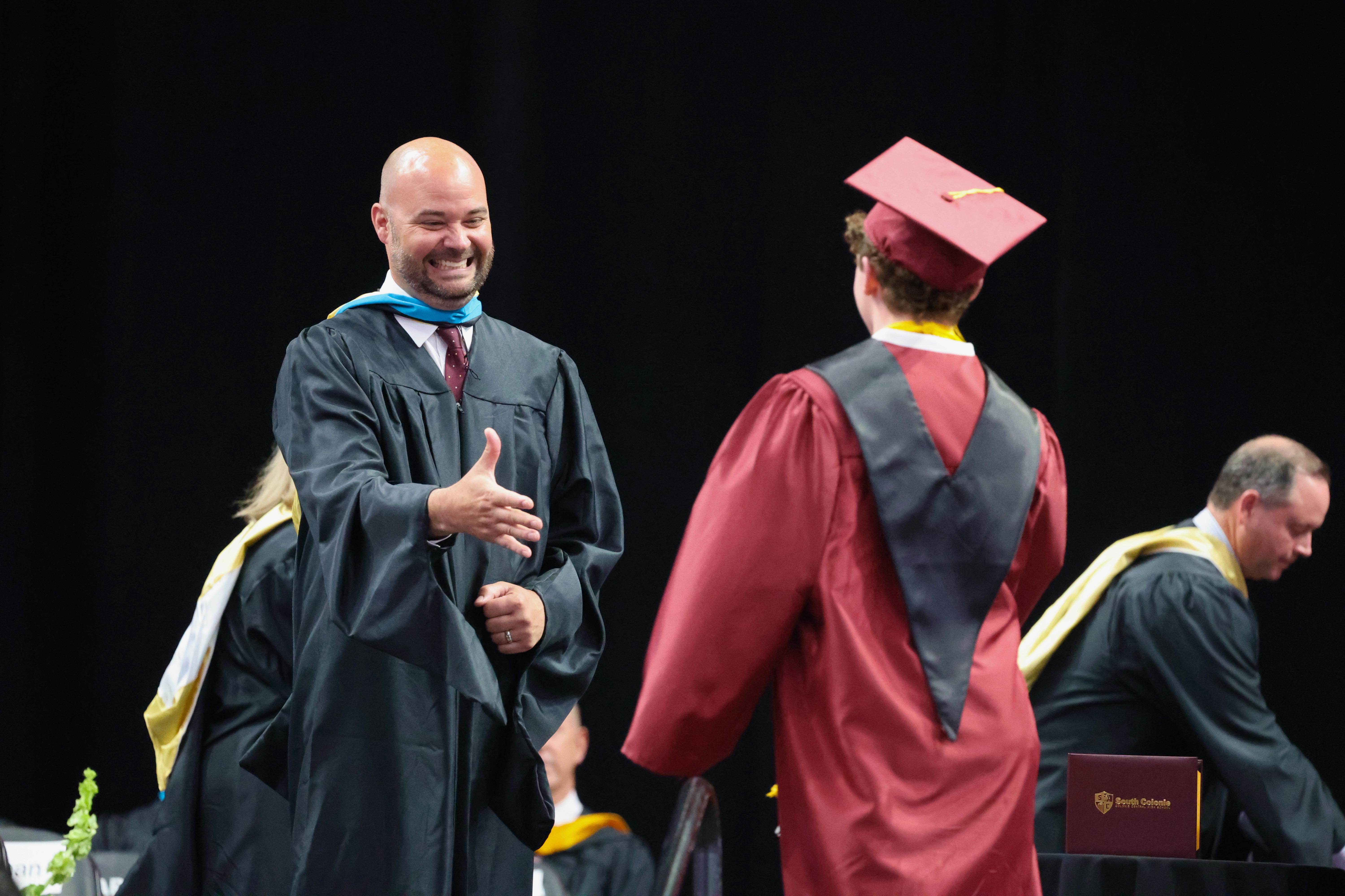 A graduate in a maroon cap and gown receives congratulations from an official at a graduation ceremony.