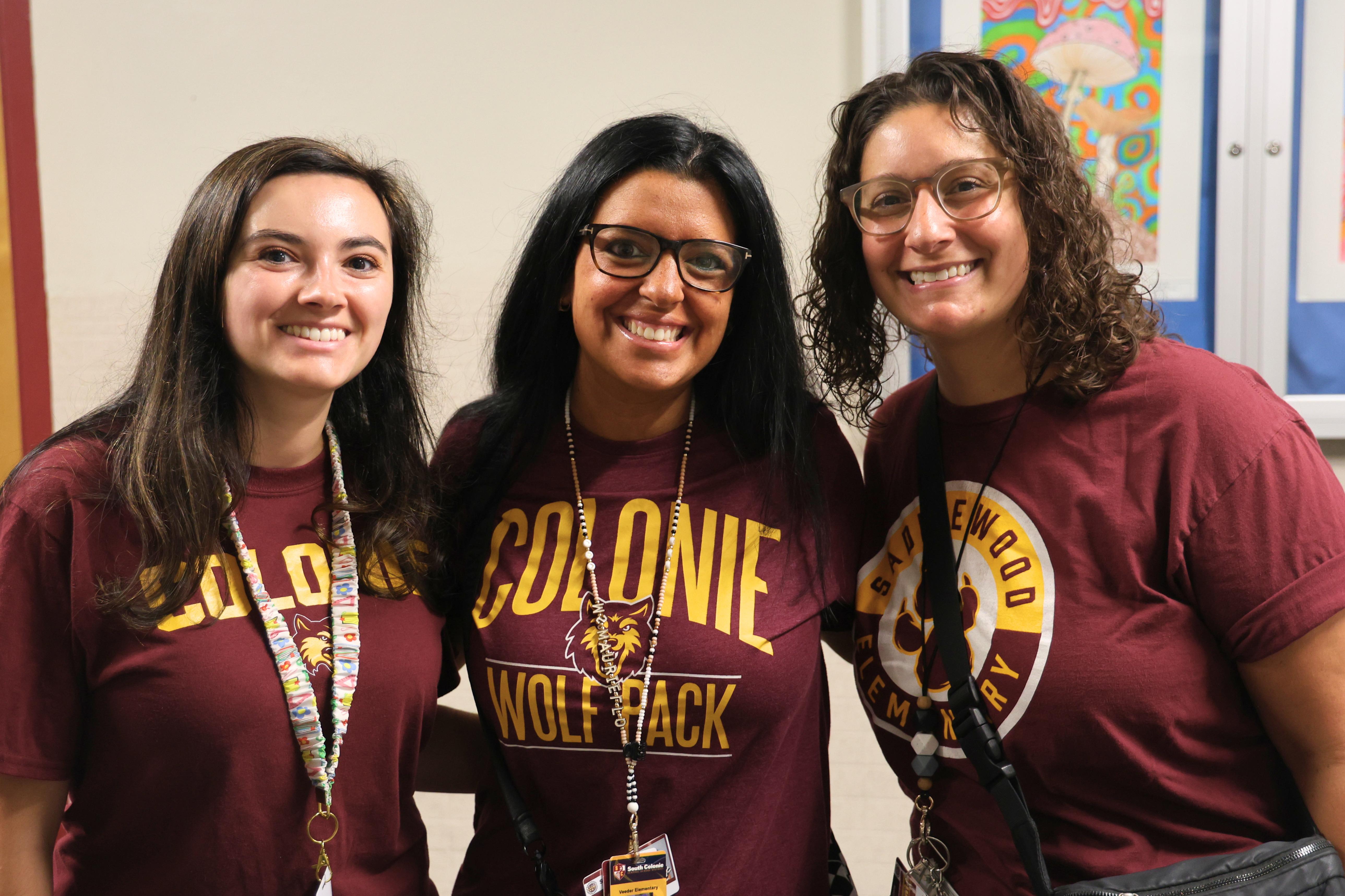Three smiling individuals pose together indoors, wearing matching maroon t-shirts that read "Colonie Wolf Pack'".