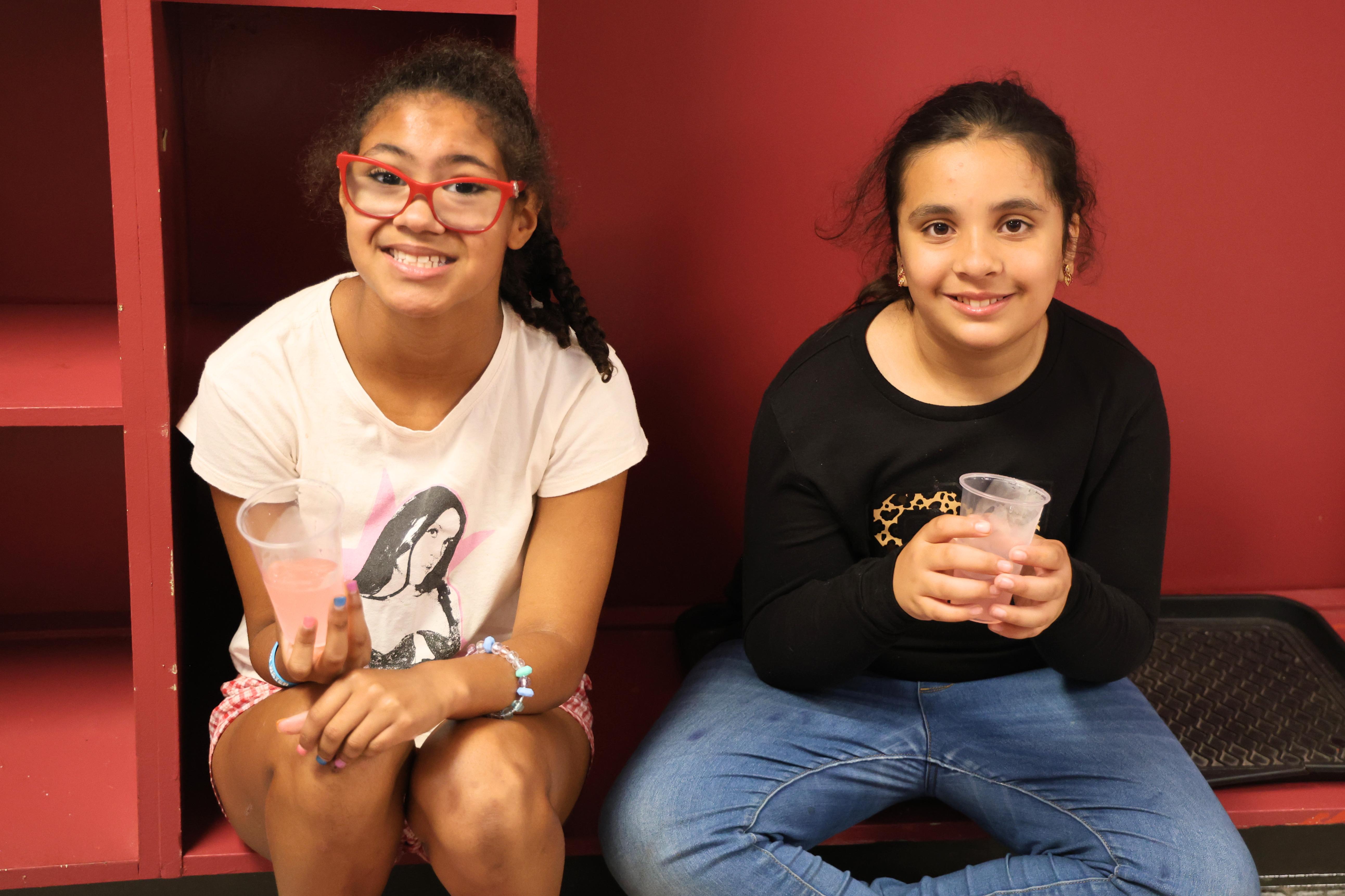 Two young girls smile at the camera, sitting side-by-side indoors holding cups of lemonade.