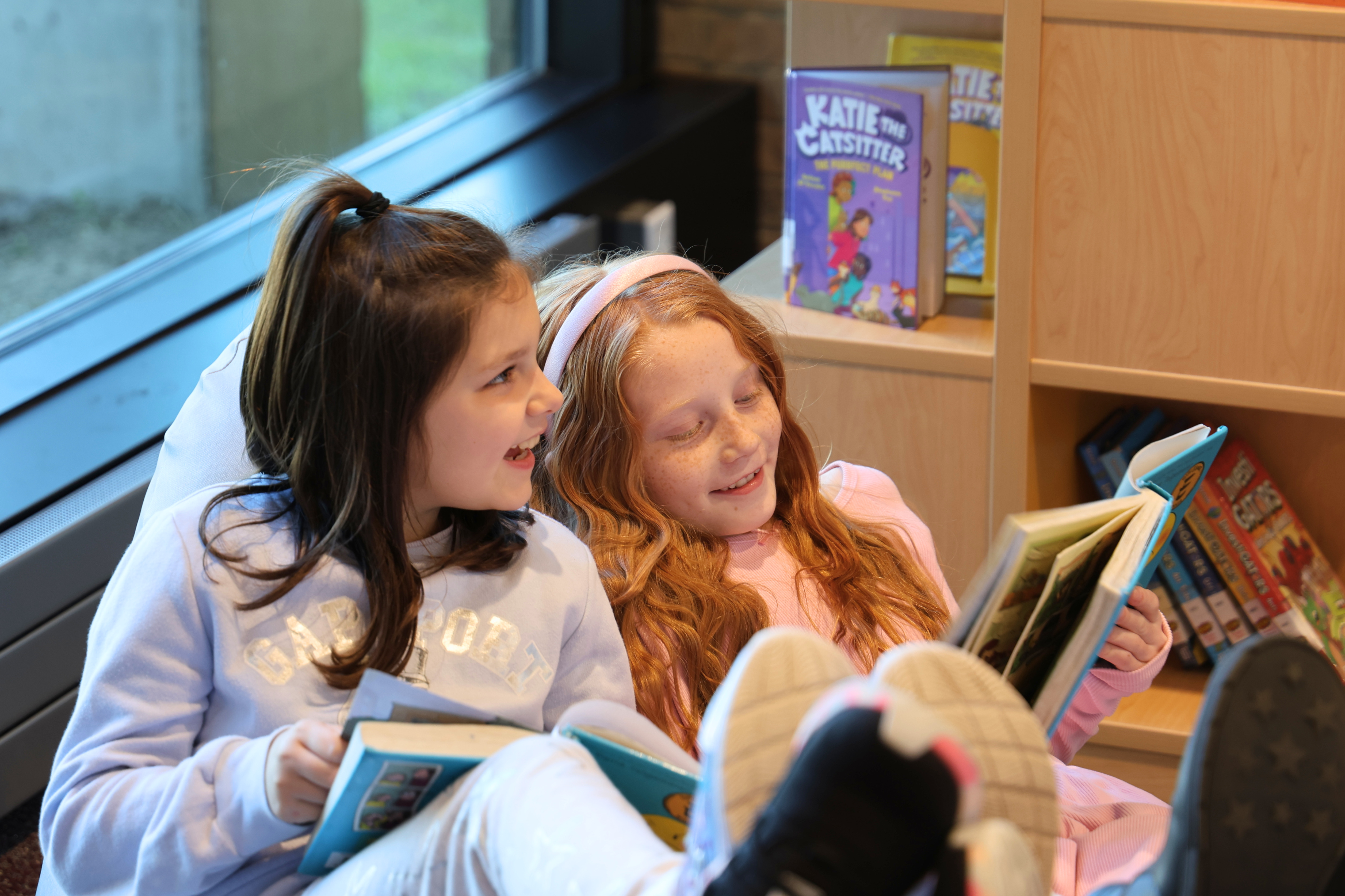 Two young girls sit comfortably indoors near a window, reading books together.