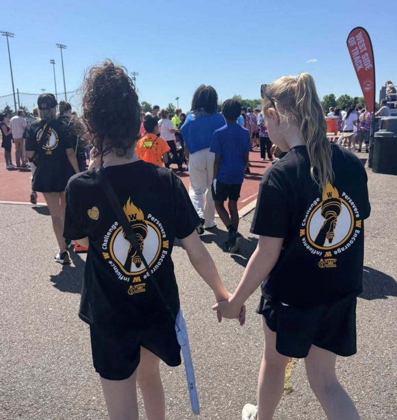 Two young girls in matching black t-shirts with a circular logo walk hand-in-hand outdoors.