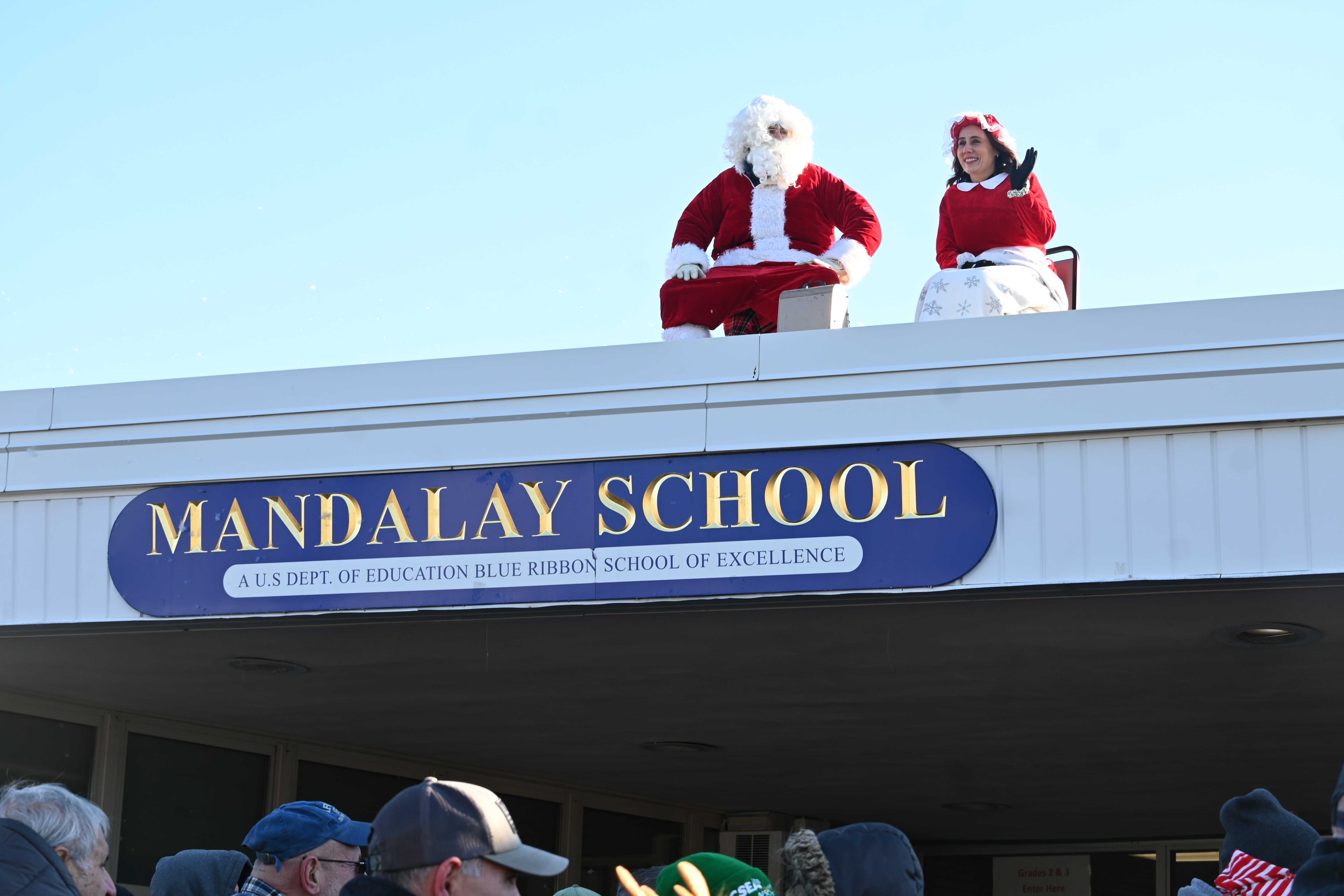 Santa Claus and Mrs. Claus sit on a rooftop above a school sign.