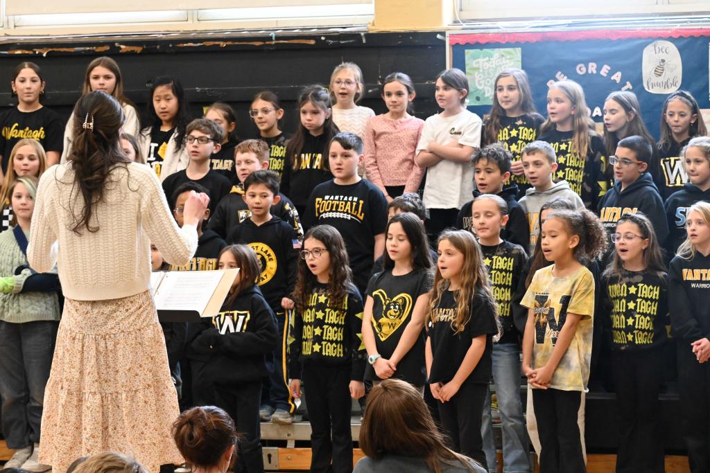 A school choir performs, led by a conductor, with students wearing matching shirts.