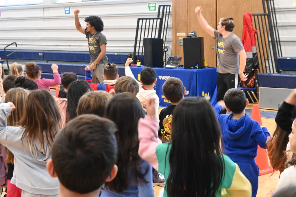 Children and adults raise their arms in a gymnasium, possibly participating in an activity.
