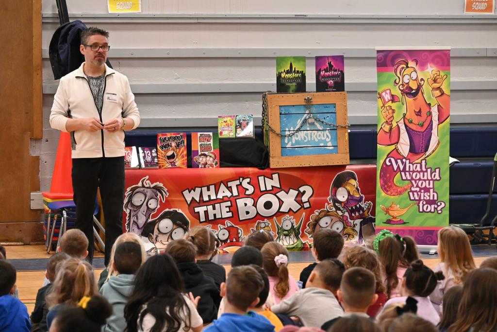 A man speaks to a group of children in front of a table with books and a sign that says 'What's in the Box?'