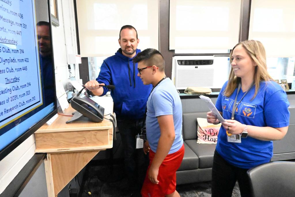 Three people stand near a desk with a phone and a screen displaying a schedule.
