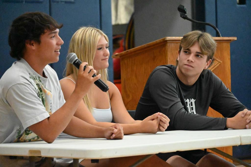 Three young people sit at a white table, with one speaking into a microphone.