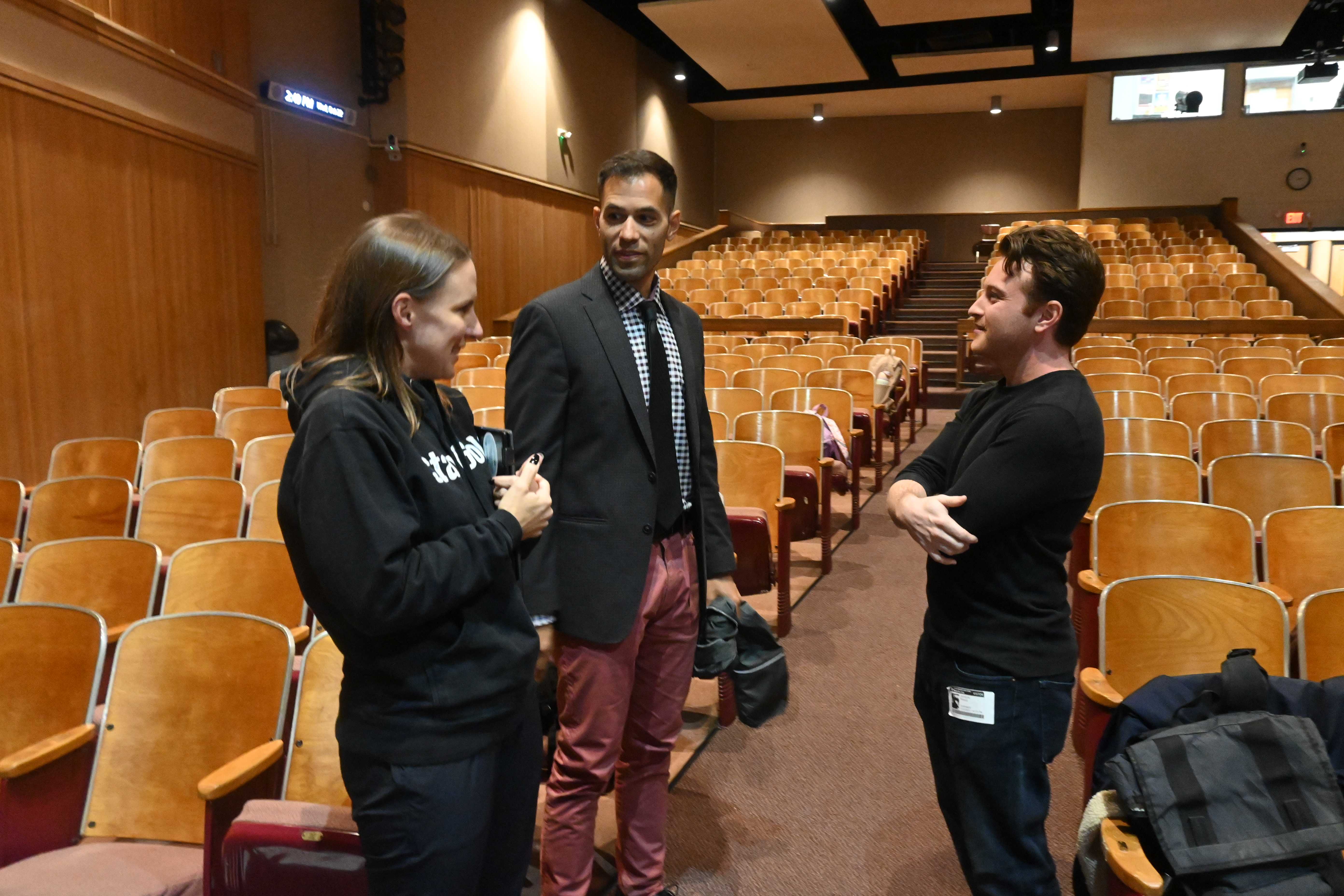Three people converse in an auditorium with rows of empty seats.
