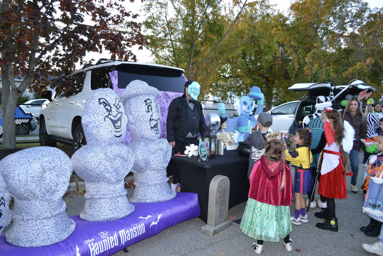 Children in costumes gather at a trunk-or-treat event, receiving treats.