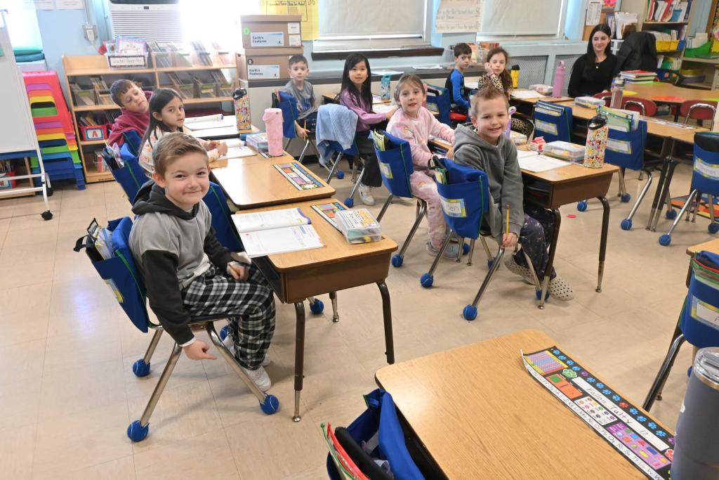Children sit at desks in a classroom, some smiling at the camera.