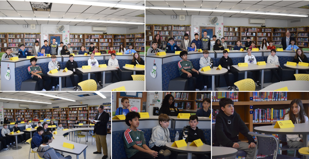 Students sit at tables in a library, some looking towards the front.