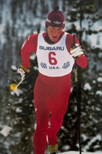 A cross-country skier, Bruce Likly, wearing a red suit with Subaru and the number six on it, and a matching hat competes in a snowy, wooded setting.