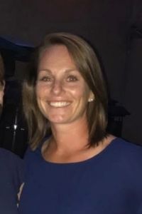 A smiling woman, Courtney Gibbons, with shoulder-length brown hair wearing a blue top against a black background.