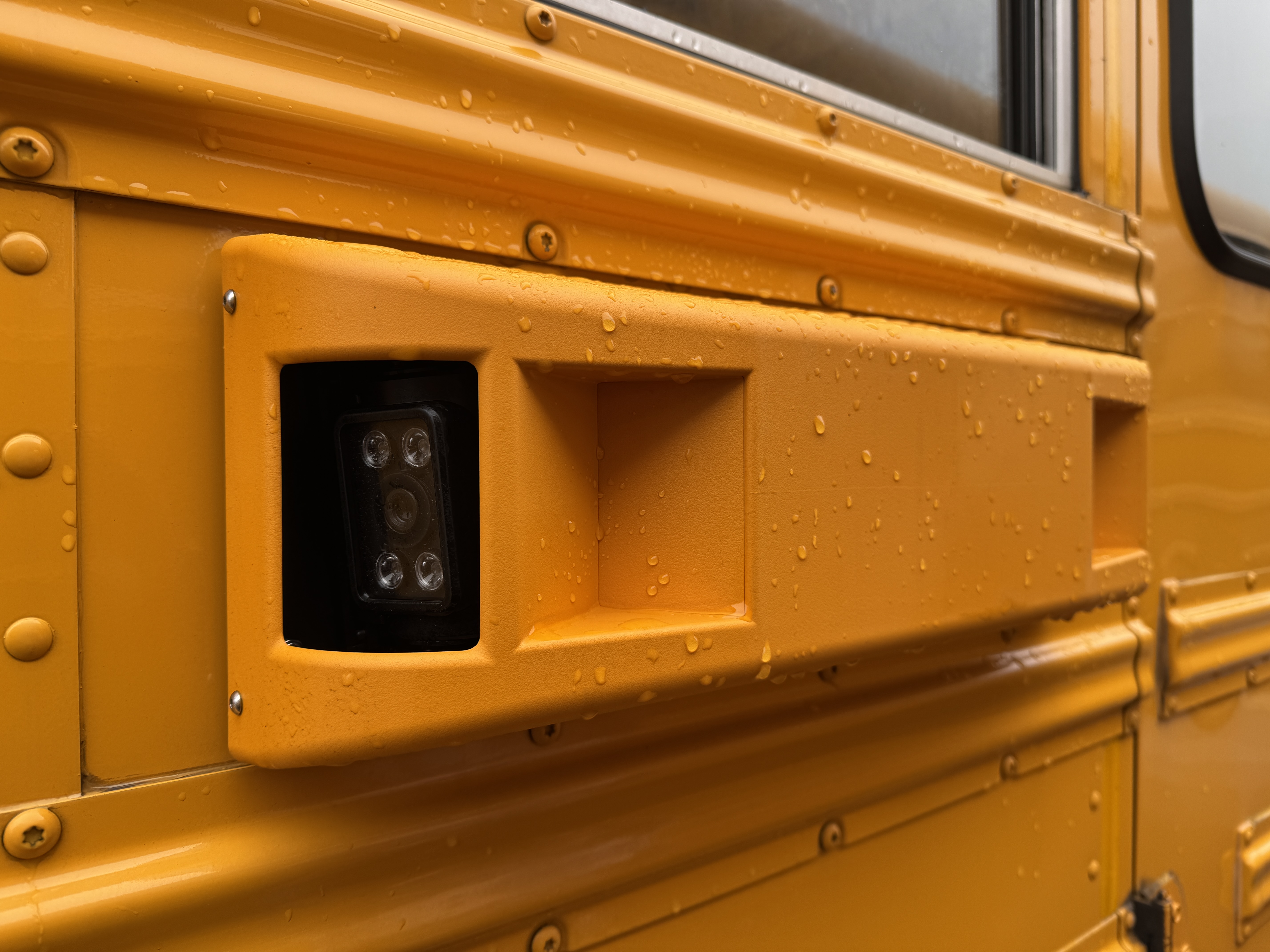 Close-up of a yellow school bus exterior with a camera and rain droplets.