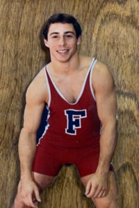 A young man, Jacques Monte, in a red wrestling singlet with the letter F on the front stands before a wooden background.