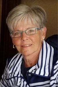 A portrait of a smiling woman, Jodi Schmeelk, with short blonde hair and glasses, wearing a blue and white striped top.