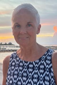 A woman, Lori Consadine, with short white hair stands outdoors wearing a blue and white patterned sleeveless top.