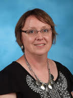 A woman with short brown hair and glasses smiles at the camera, wearing a black top with a decorative neckline.