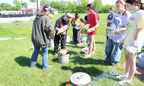 Two students use tongs to dip a piece of art in a Raku kiln, while fellow students look on.