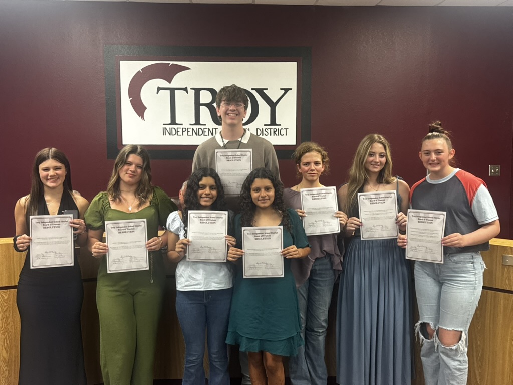 A group of students hold certificates in front of a 'Troy' sign.