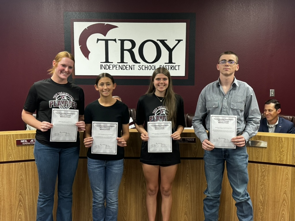 Four people stand holding certificates in front of a 'TROY' sign.