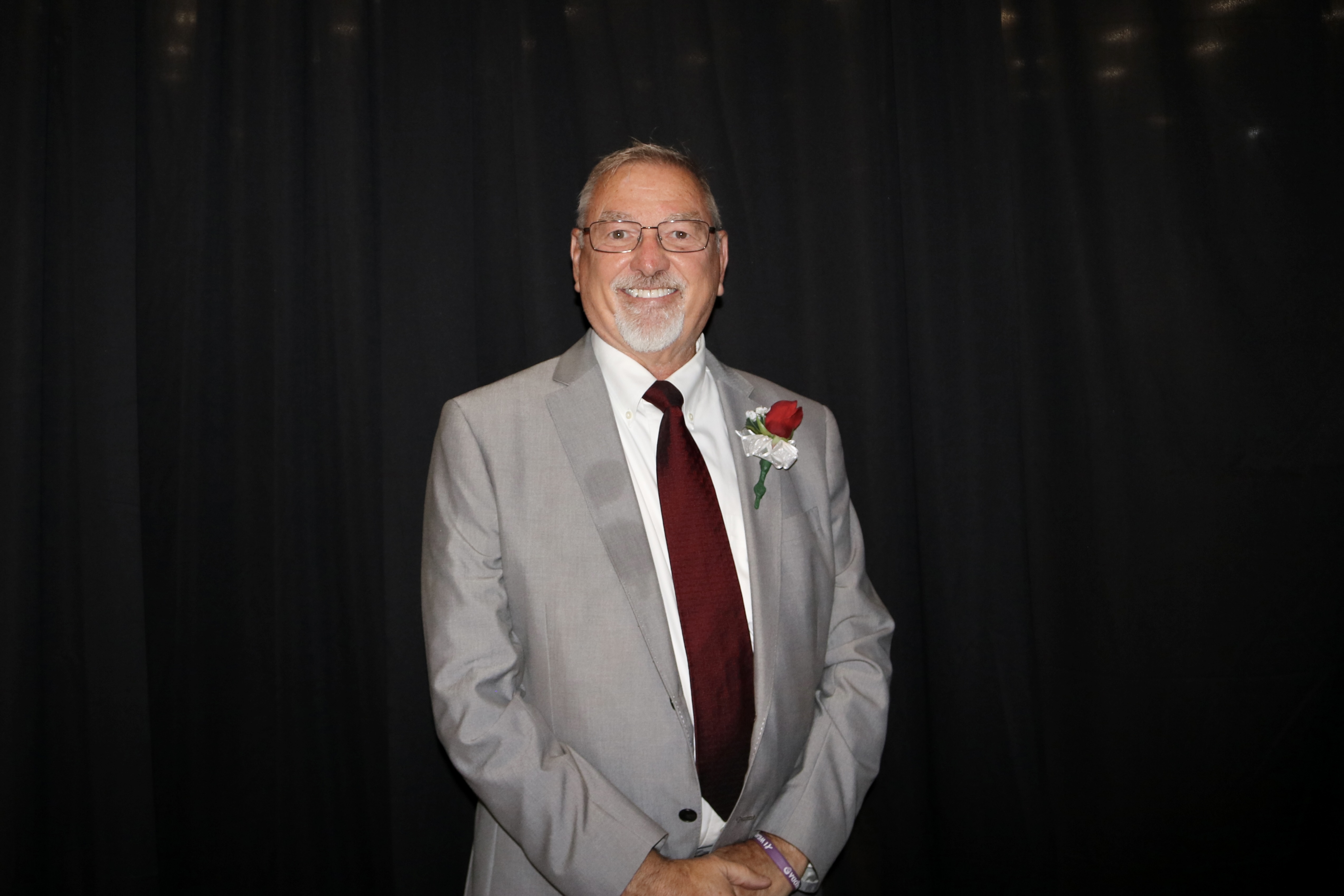 A smiling man in a light gray suit and burgundy tie stands against a black backdrop.