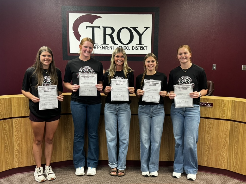 Five young women stand together, holding certificates in front of a 'Troy' sign.