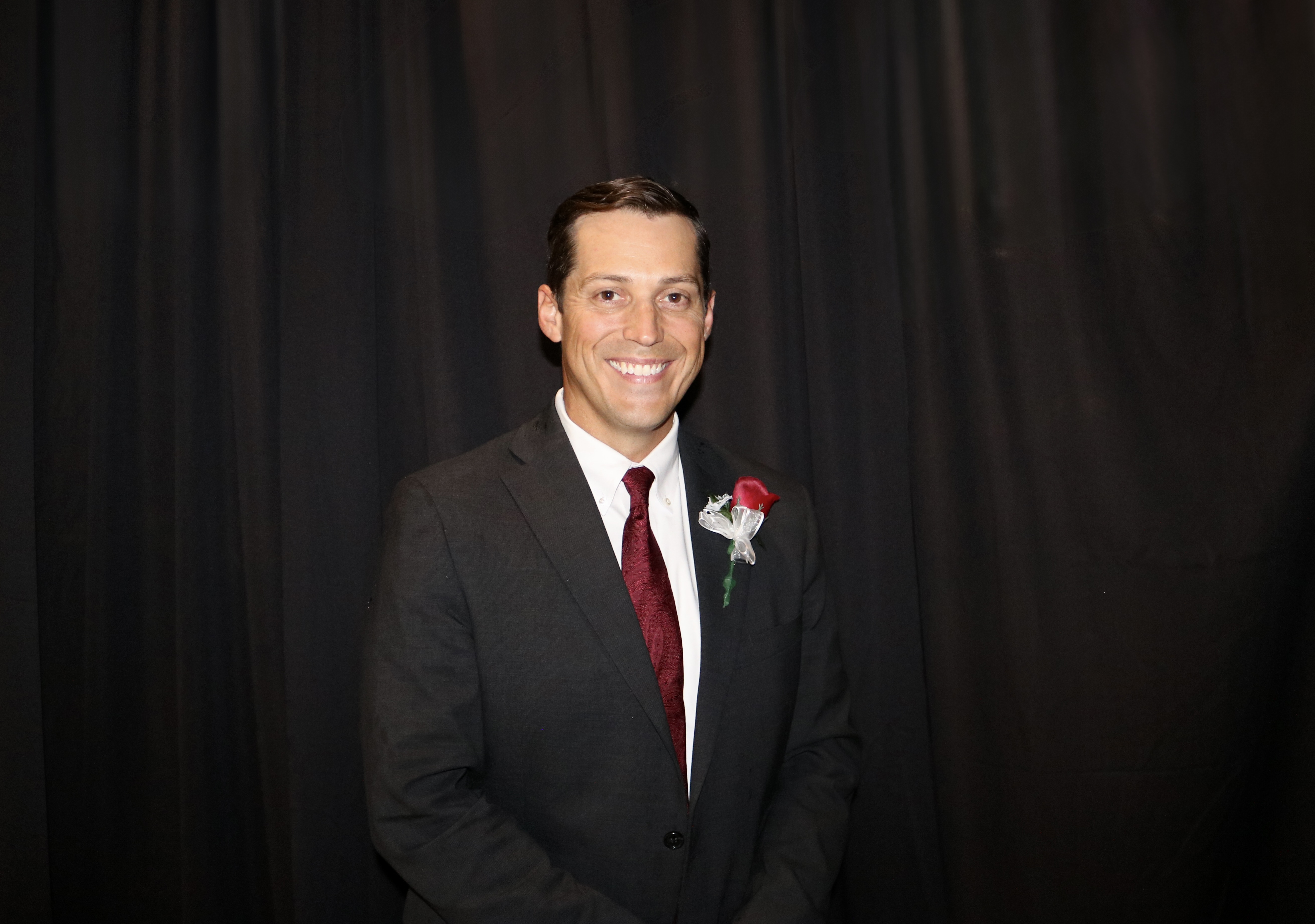 A man in a dark suit and red tie smiles at the camera against a black backdrop.