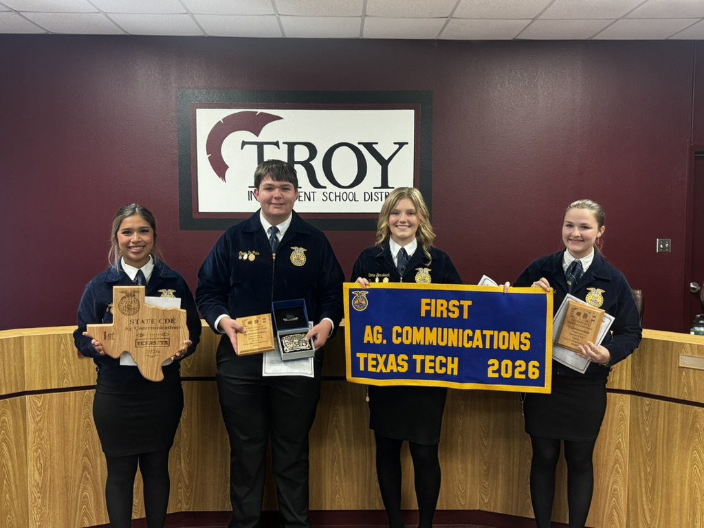Four students in uniform hold awards, smiling in front of a 'Troy' sign.