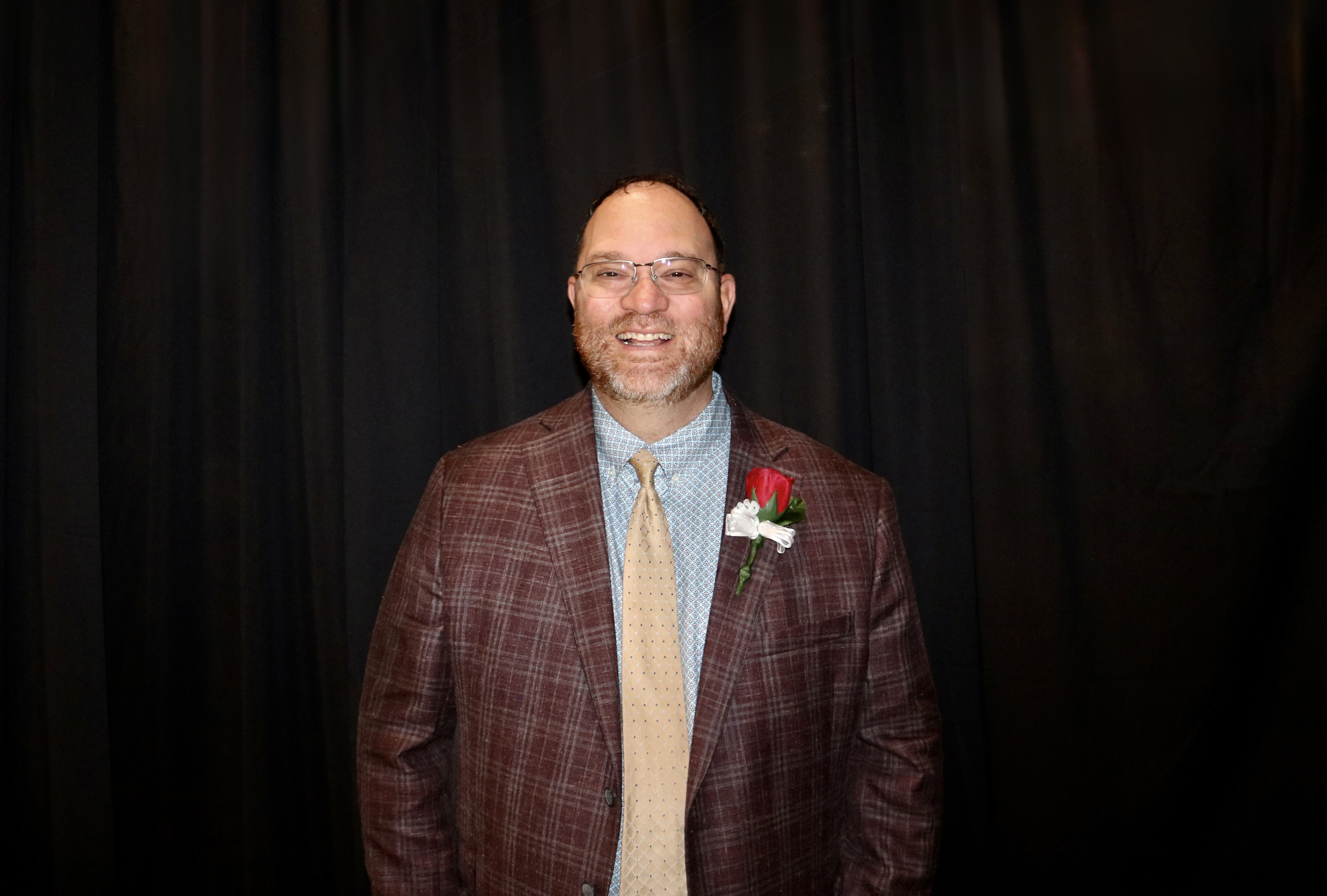 A man in a patterned jacket and tie smiles at the camera against a dark backdrop.