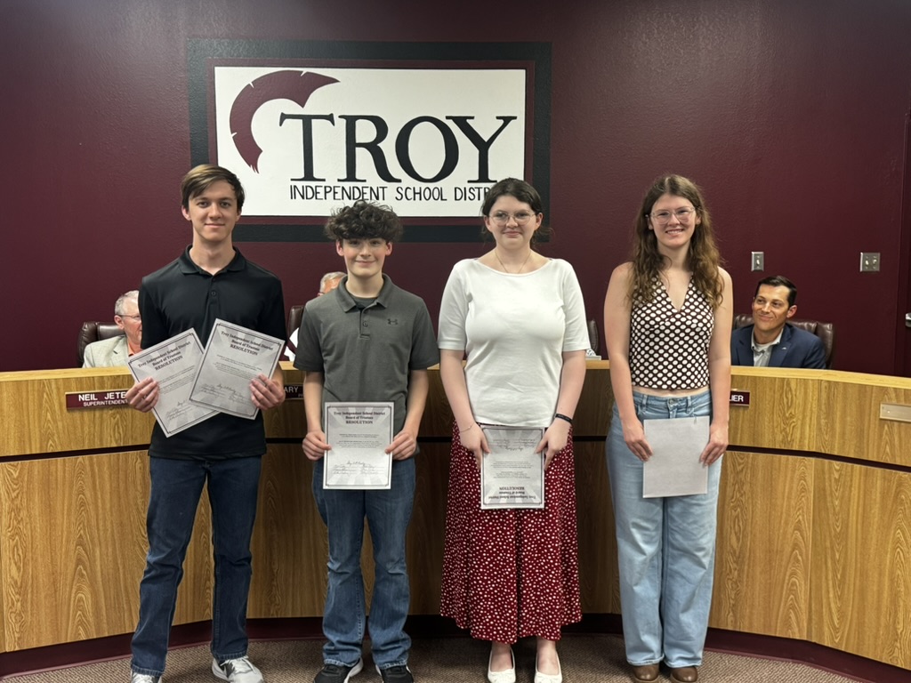 Four people stand holding papers in front of a 'TROY' sign.