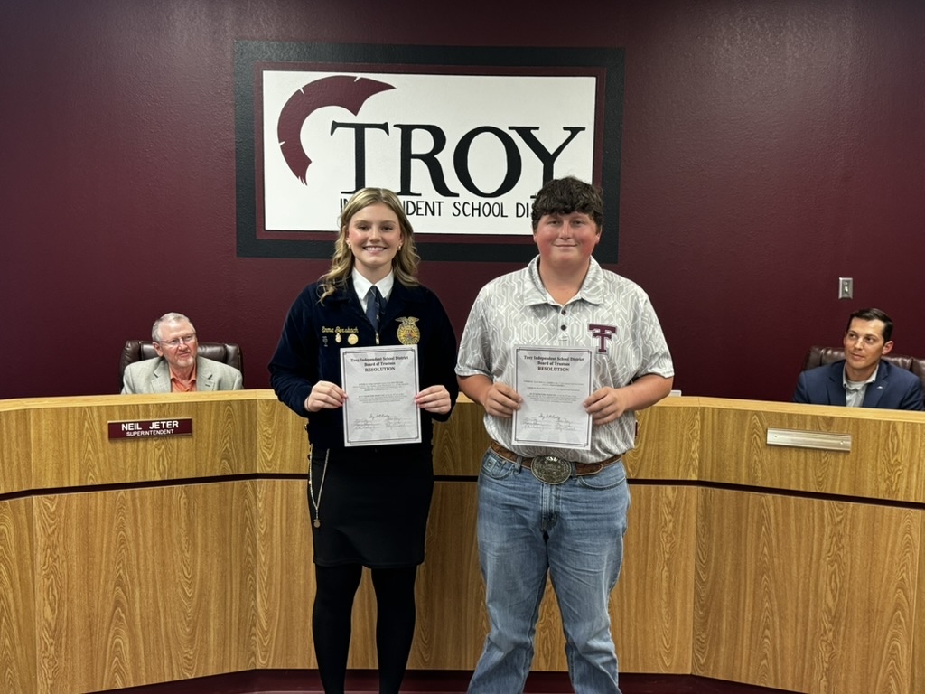 Two people stand holding certificates in front of a 'Troy' sign.
